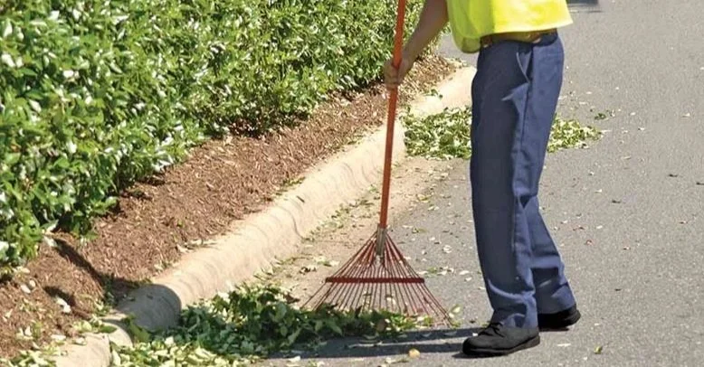 A person wearing a yellow safety vest and navy pants raking leaves or debris along the edge of a sidewalk.