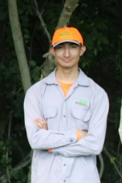 A young man standing outdoors with arms crossed, wearing a gray shirt with orange accents and a bright orange cap, in front of a green, wooded background.