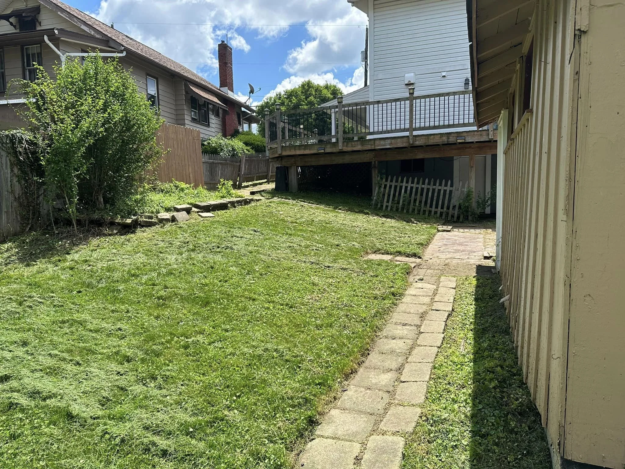 A backyard with green grass, a stone pathway, a bush on the left, and a wooden deck attached to a house on the right. The sky is partly cloudy.