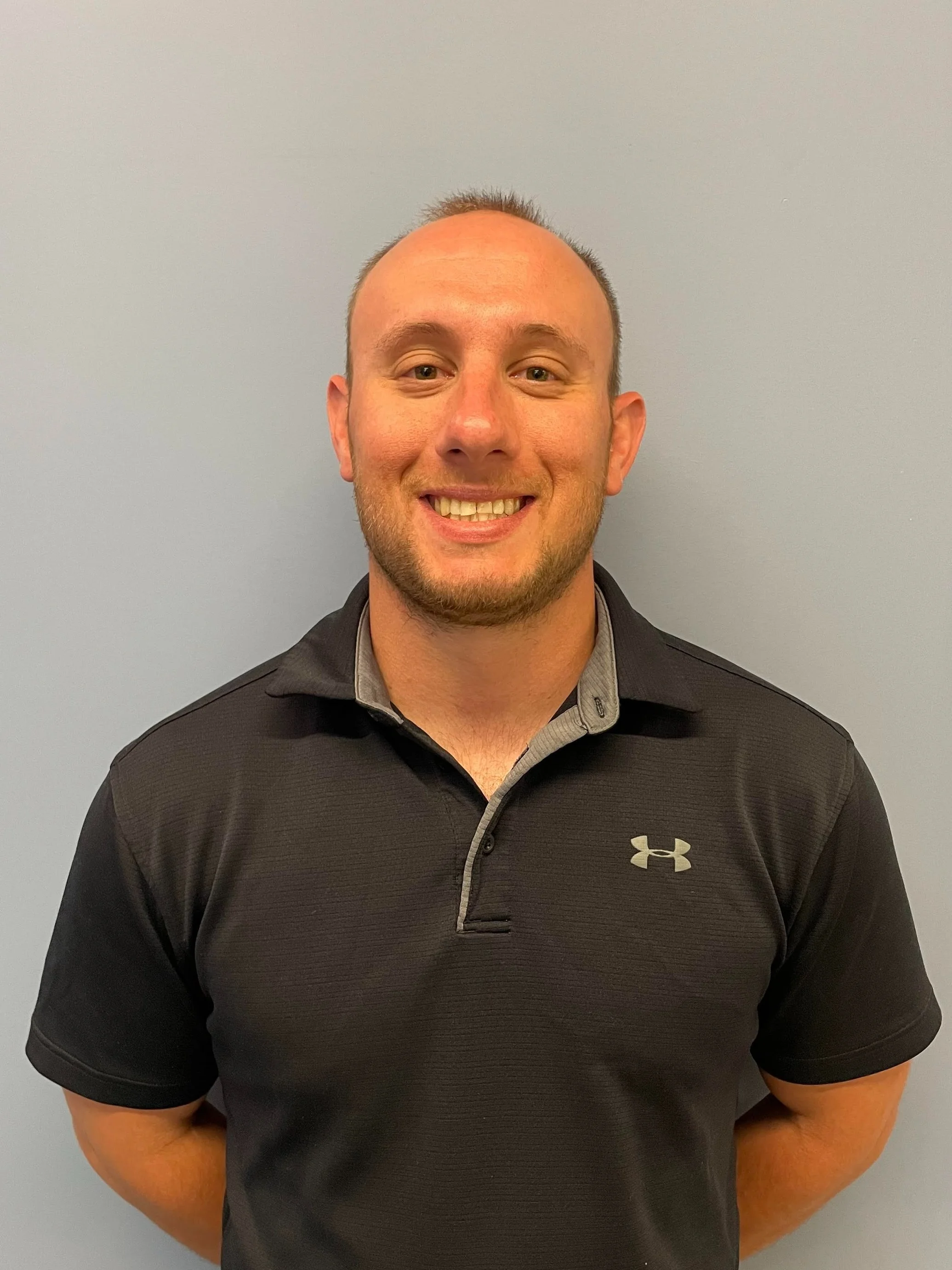 Man in a black polo shirt standing against a light-colored wall, smiling.