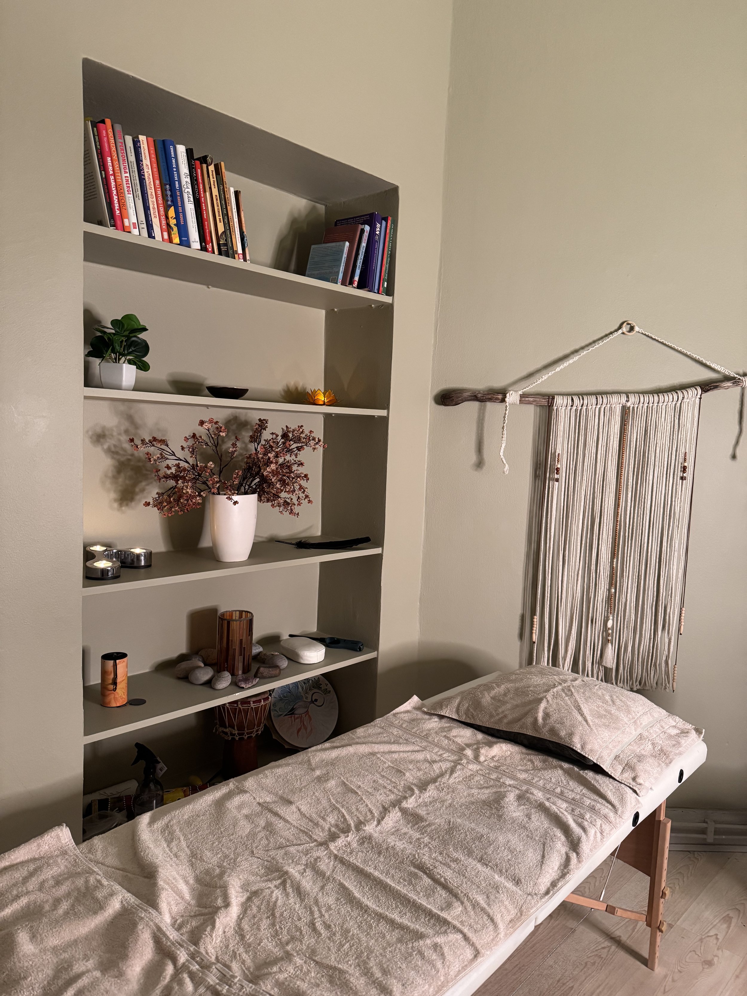 Säng med beige sängkläder och två kuddar framför ett vitt hyllsystem med växter och ljuslyktor, samt en väggbonad i växtstil.