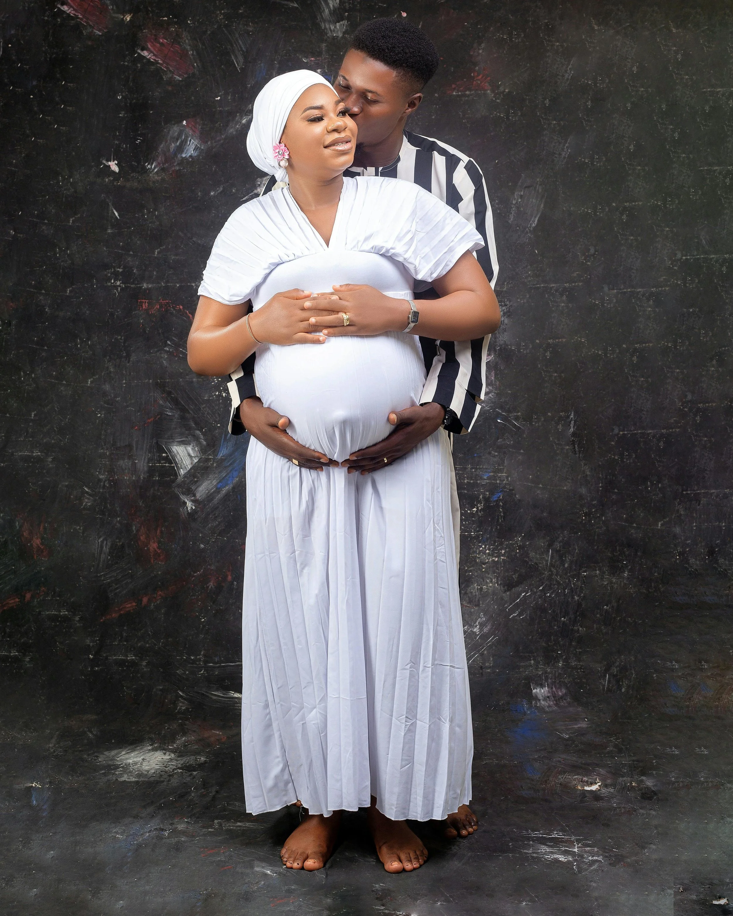 A pregnant woman in white dress and headwrap stands with a man behind her, who is kissing her on the forehead; they are both holding her pregnant belly, against a dark textured background.