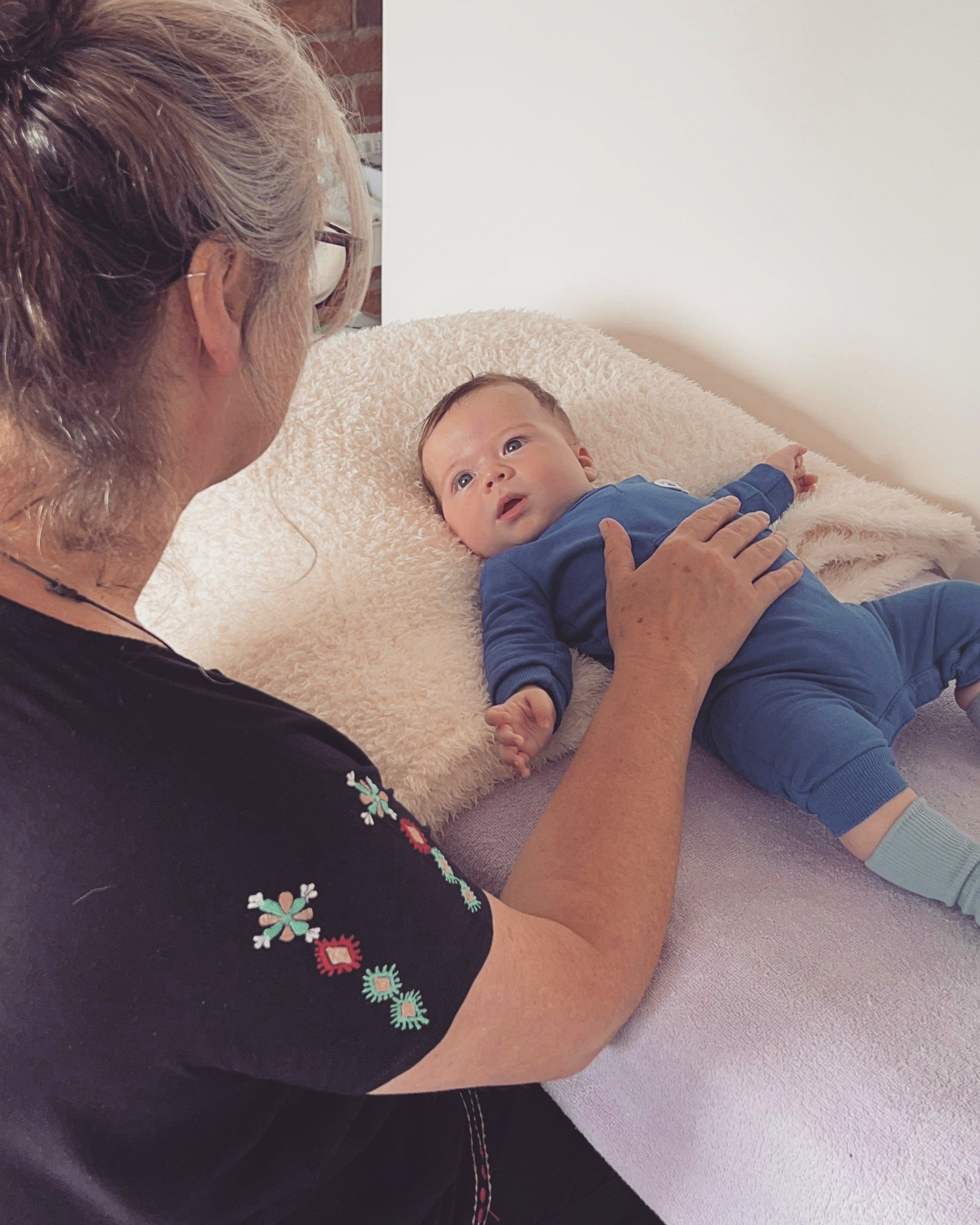 An elderly woman with glasses and embroidered black shirt touching a baby lying on a bed, looking at her.