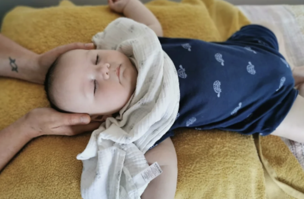 A young child with closed eyes lying on a yellow blanket receiving a gentle head massage from an adult with two hands.