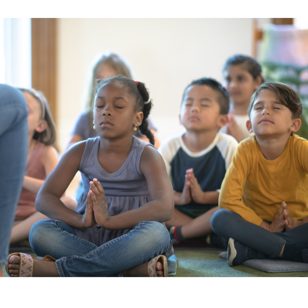 Children meditating with eyes closed, sitting cross-legged on the floor in a classroom or meditation space.