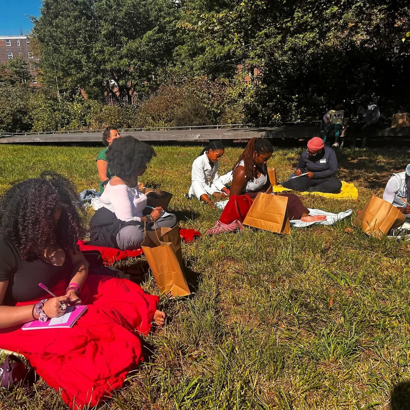 Group of women sitting on the grass in a park, some writing in notebooks, with brown paper bags around them, under trees with a city building in the background.