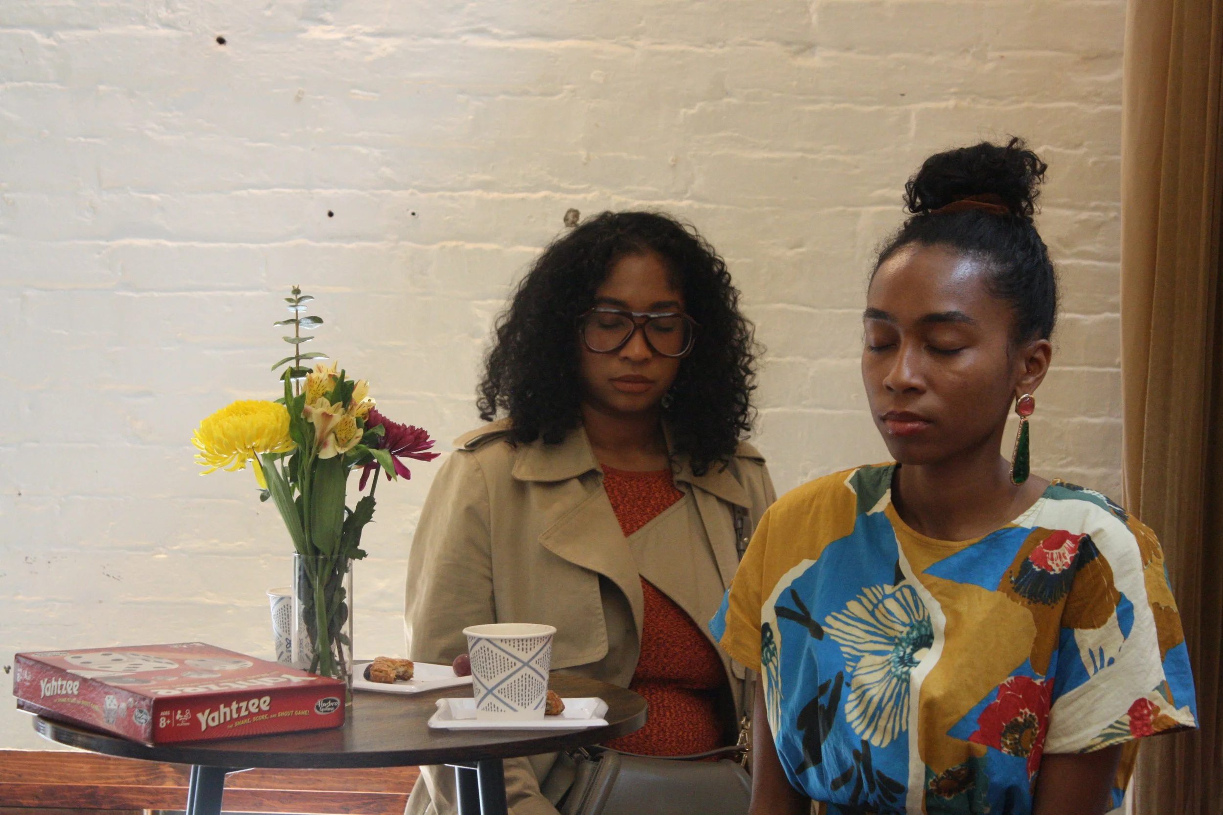 Two women with closed eyes sitting at a table with flowers, food, and game box in front of them against a white brick wall.