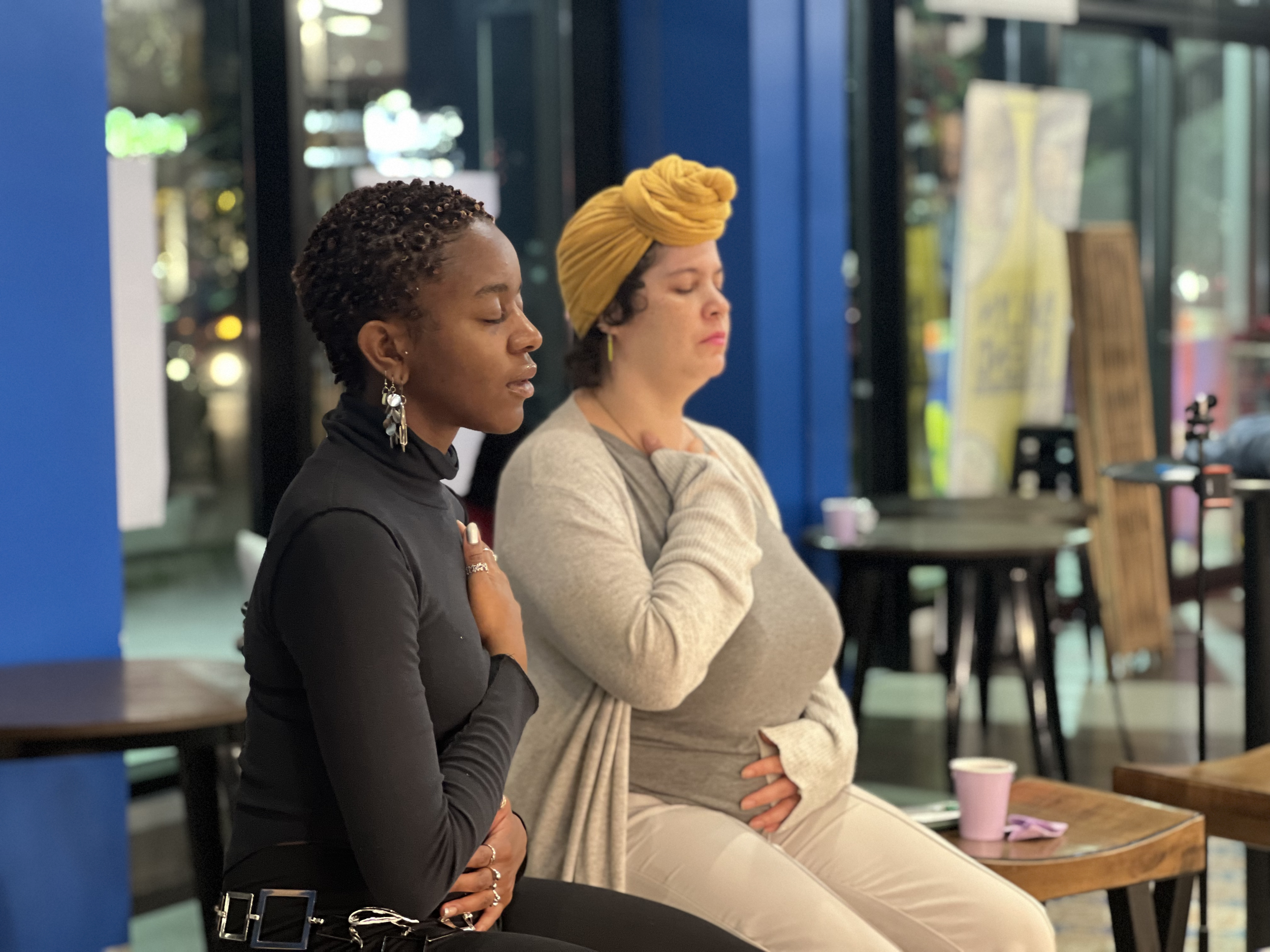 Two women sitting with eyes closed, meditating in a modern indoor space, with one woman wearing a yellow head wrap, and the other in black top and earrings.
