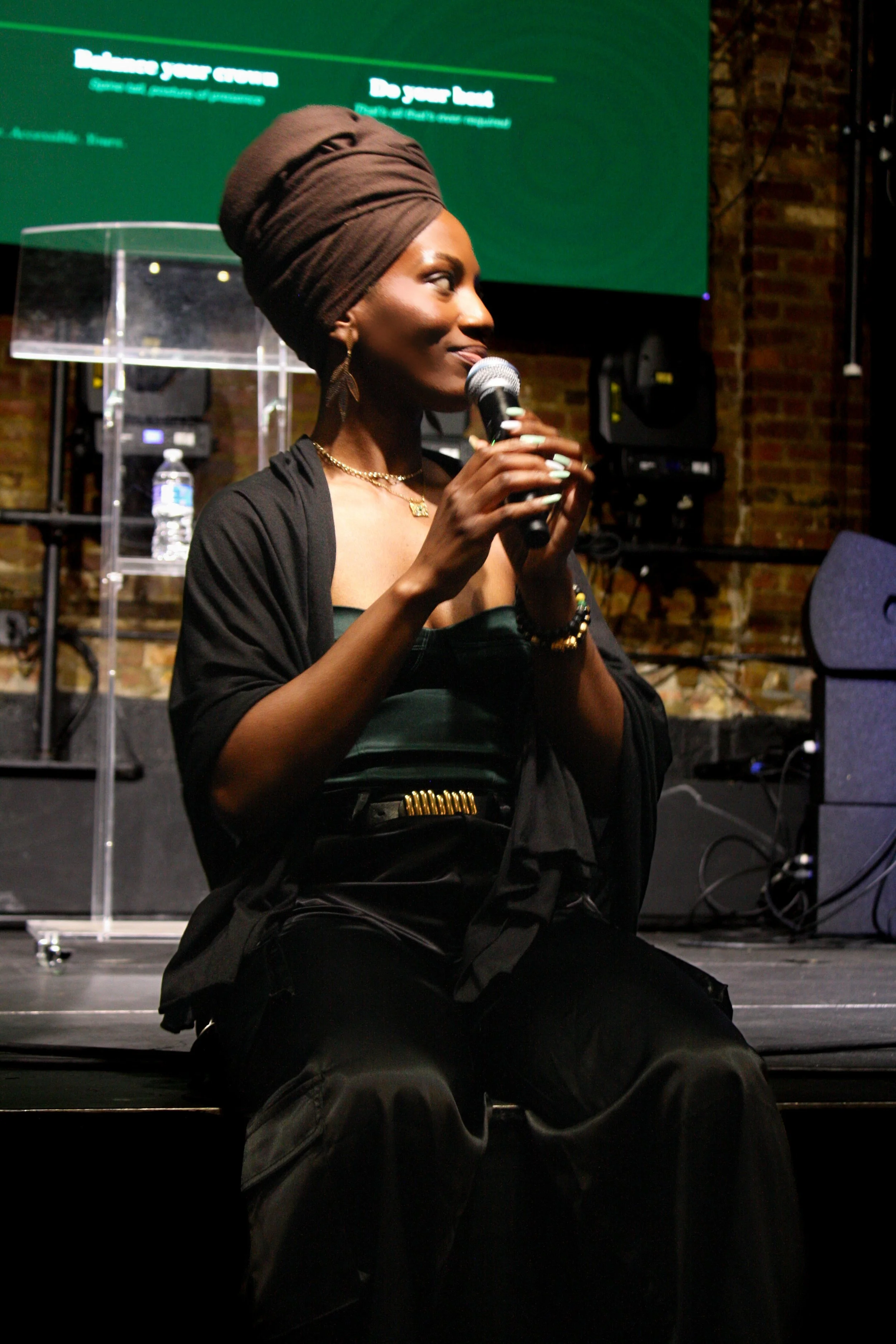 A woman wearing a brown headwrap and black clothing speaking into a microphone on stage with a green screen and brick wall background.