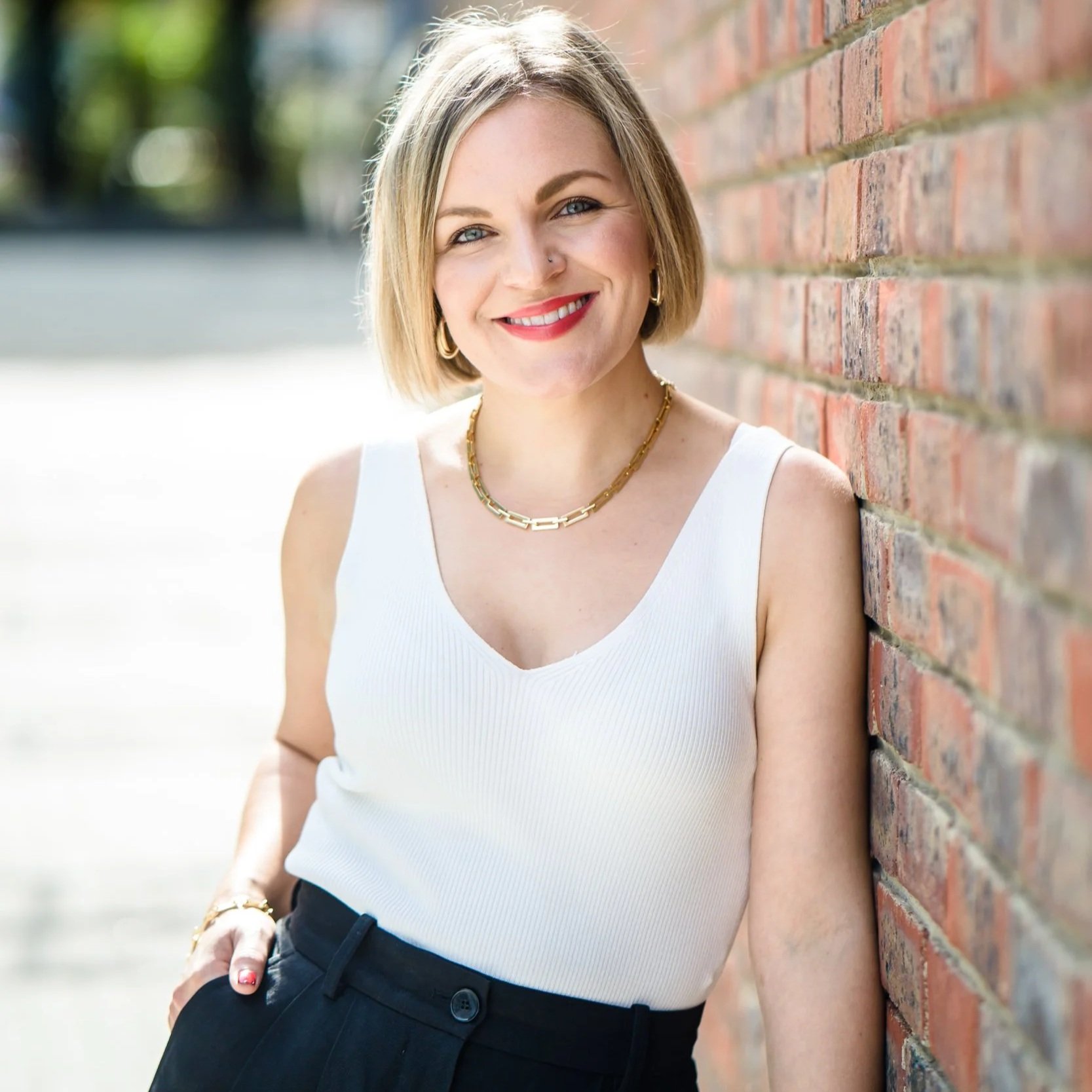 A woman with blonde hair smiling, wearing a white sleeveless top, gold necklace, earrings, and a bracelet, standing next to a brick wall outdoors.