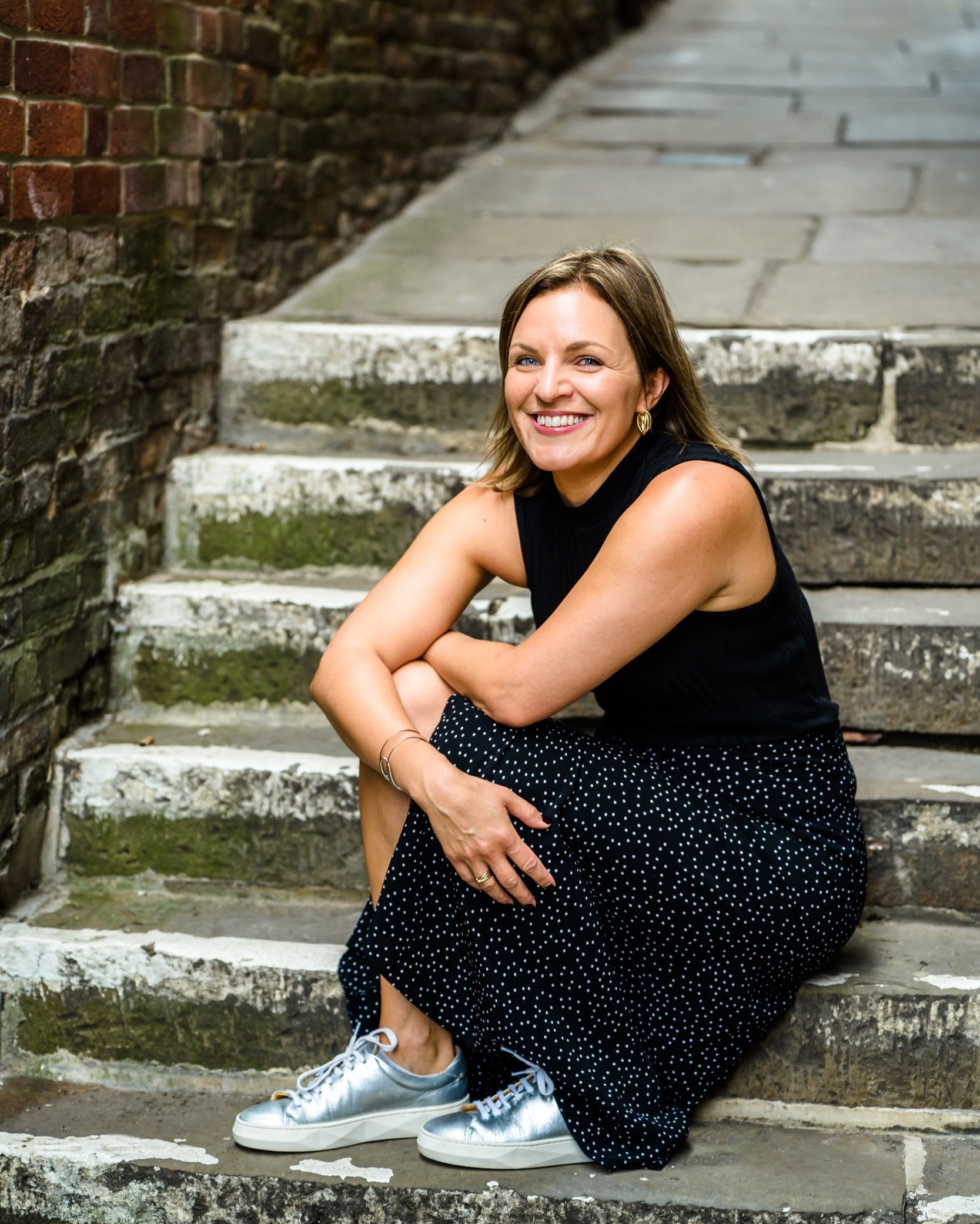 Woman sitting on stone steps outdoors, wearing a black sleeveless top, black polka dot skirt, white sneakers, and gold earrings, smiling at the camera.
