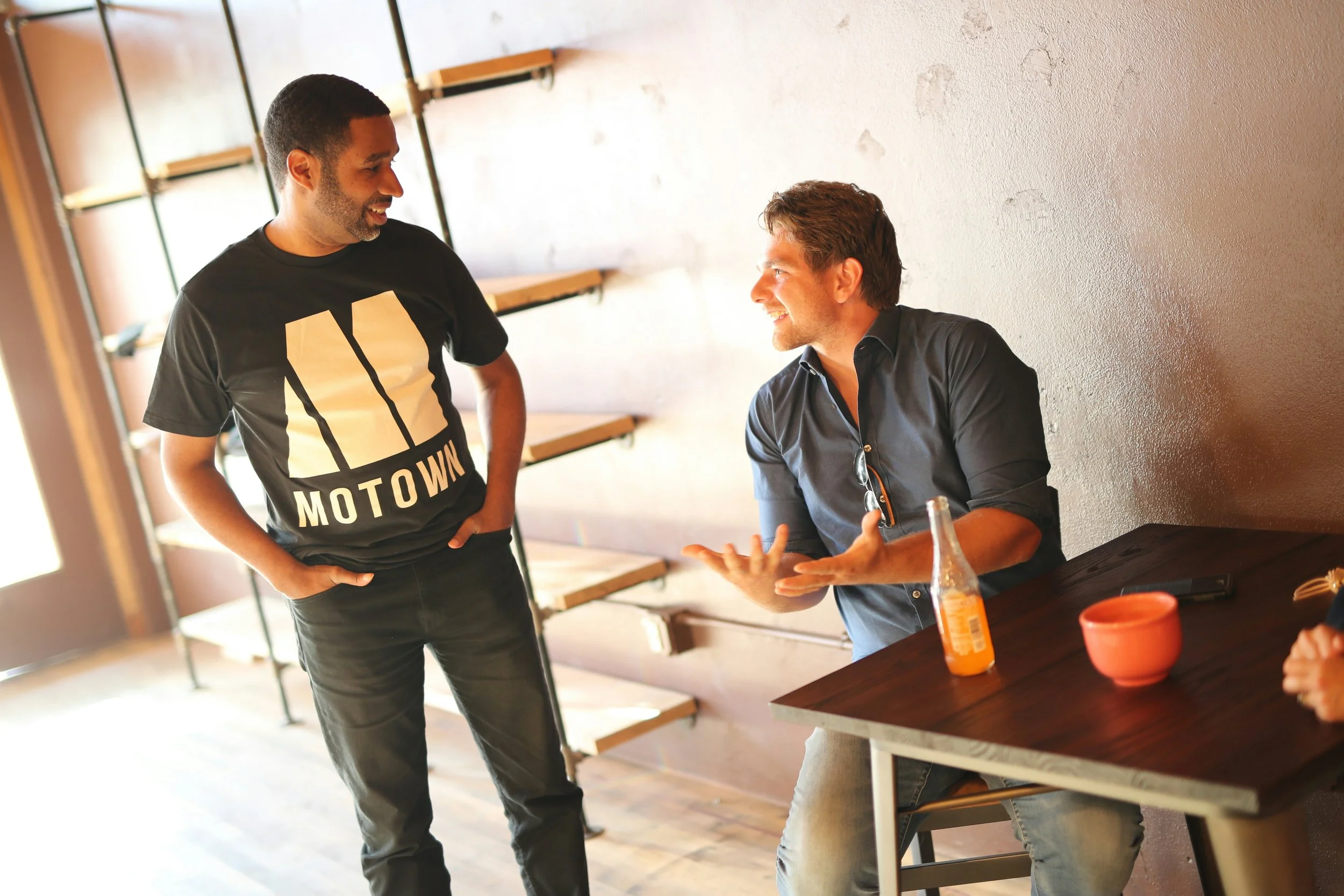 Two men having a conversation in a cafe, one standing and one sitting at a table, with a bottle and snacks on the table.