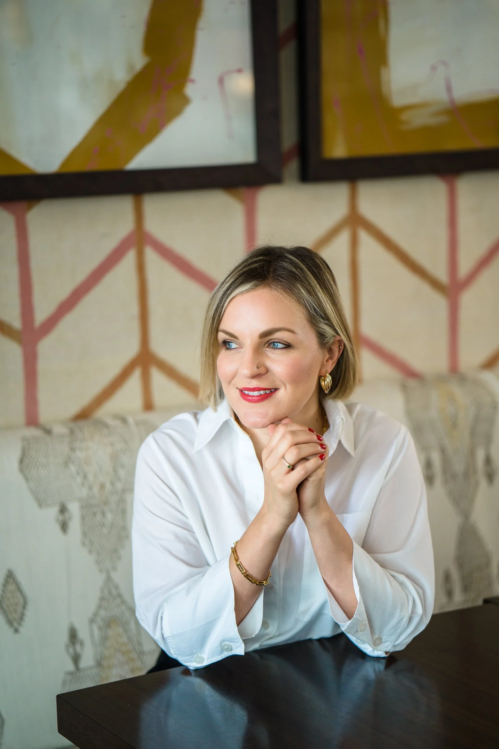 A woman with blonde hair, blue eyes, and red lipstick, wearing a white shirt and gold jewelry, sitting at a dark wooden table and smiling with her hands clasped together under her chin. The background features a muted yellow wall with colorful geometric patterns and framed artwork.