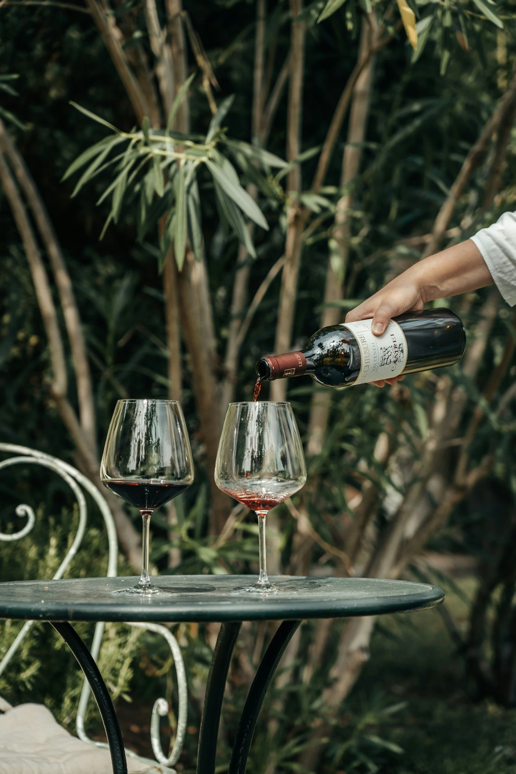 Person pouring red wine into a glass on an outdoor table with another glass of wine already filled, set against a background of leafy green plants.