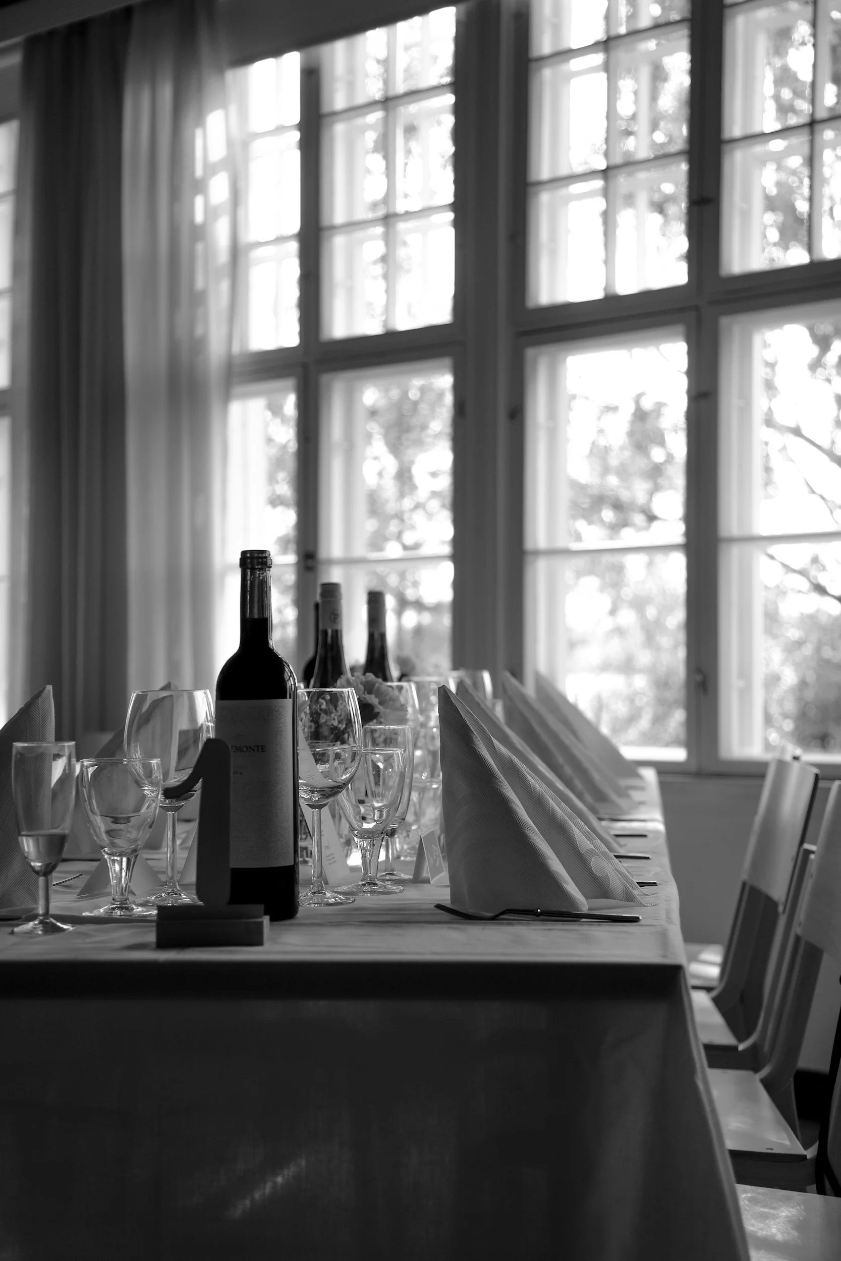 Black and white photo of a dining table set with wine bottles, glasses, and folded napkins, near large windows with curtains.