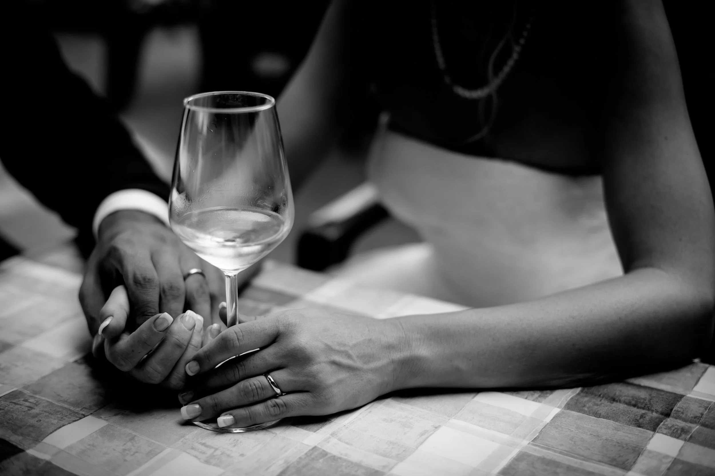 Close-up of a couple's hands on a checkered tablecloth, with the woman's hand resting on the man's hand and both wearing rings, and a wine glass held by the man's hand. The woman is wearing a dress and necklaces.