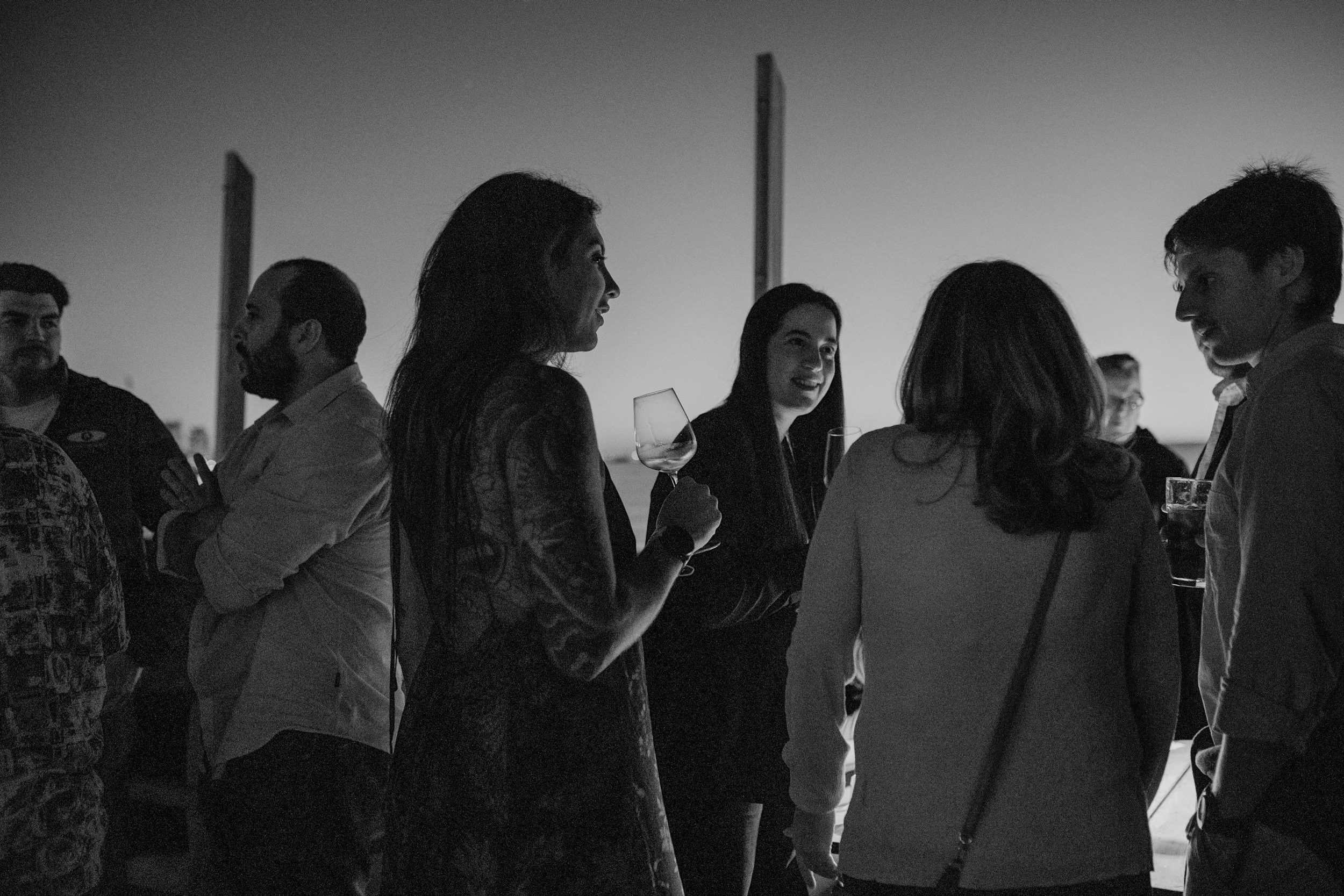 A group of people socializing at an outdoor gathering during sunset, some holding wine glasses, with a mostly clear sky in the background.