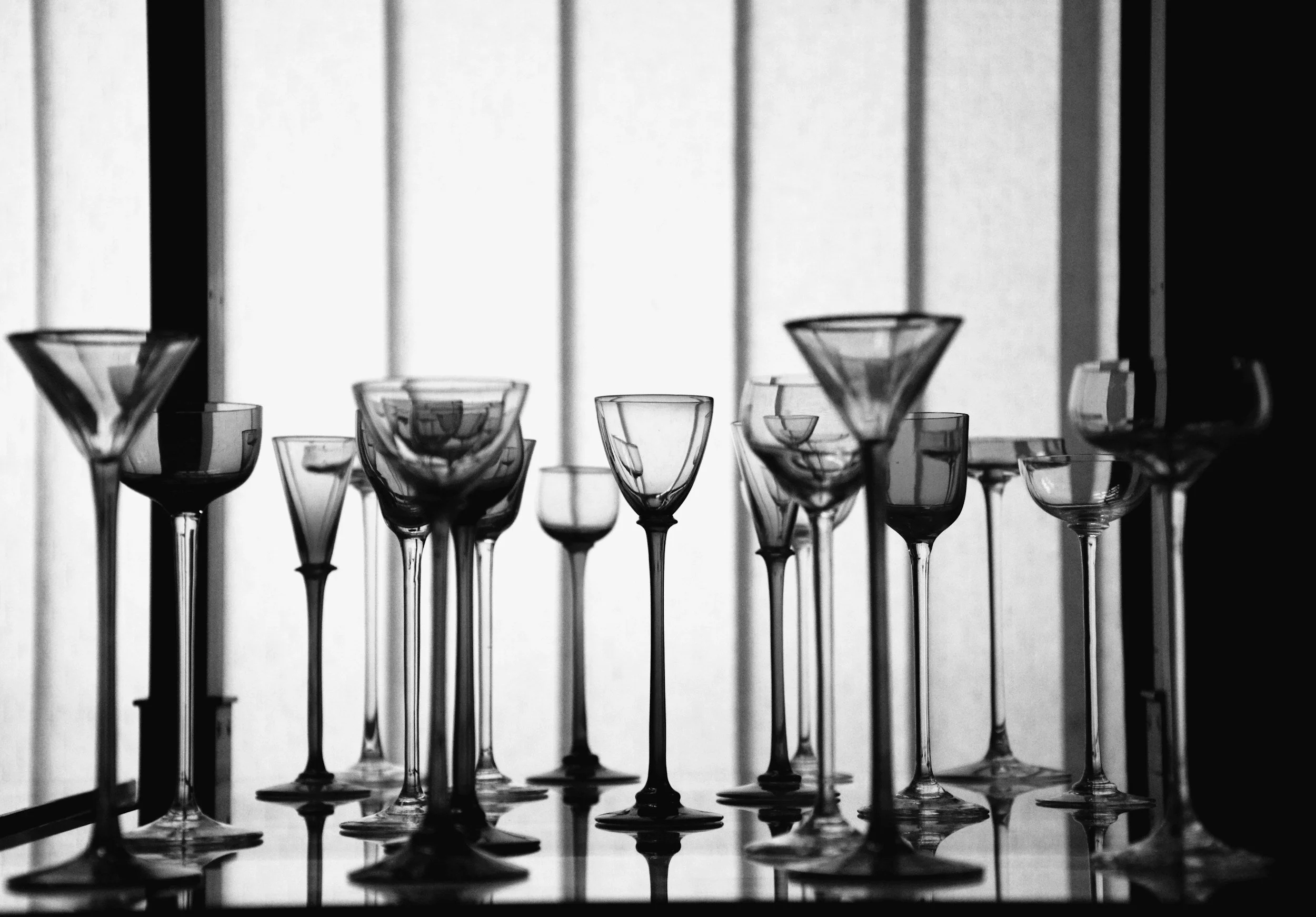 A collection of empty variously shaped glasses arranged on a reflective surface with a window in the background