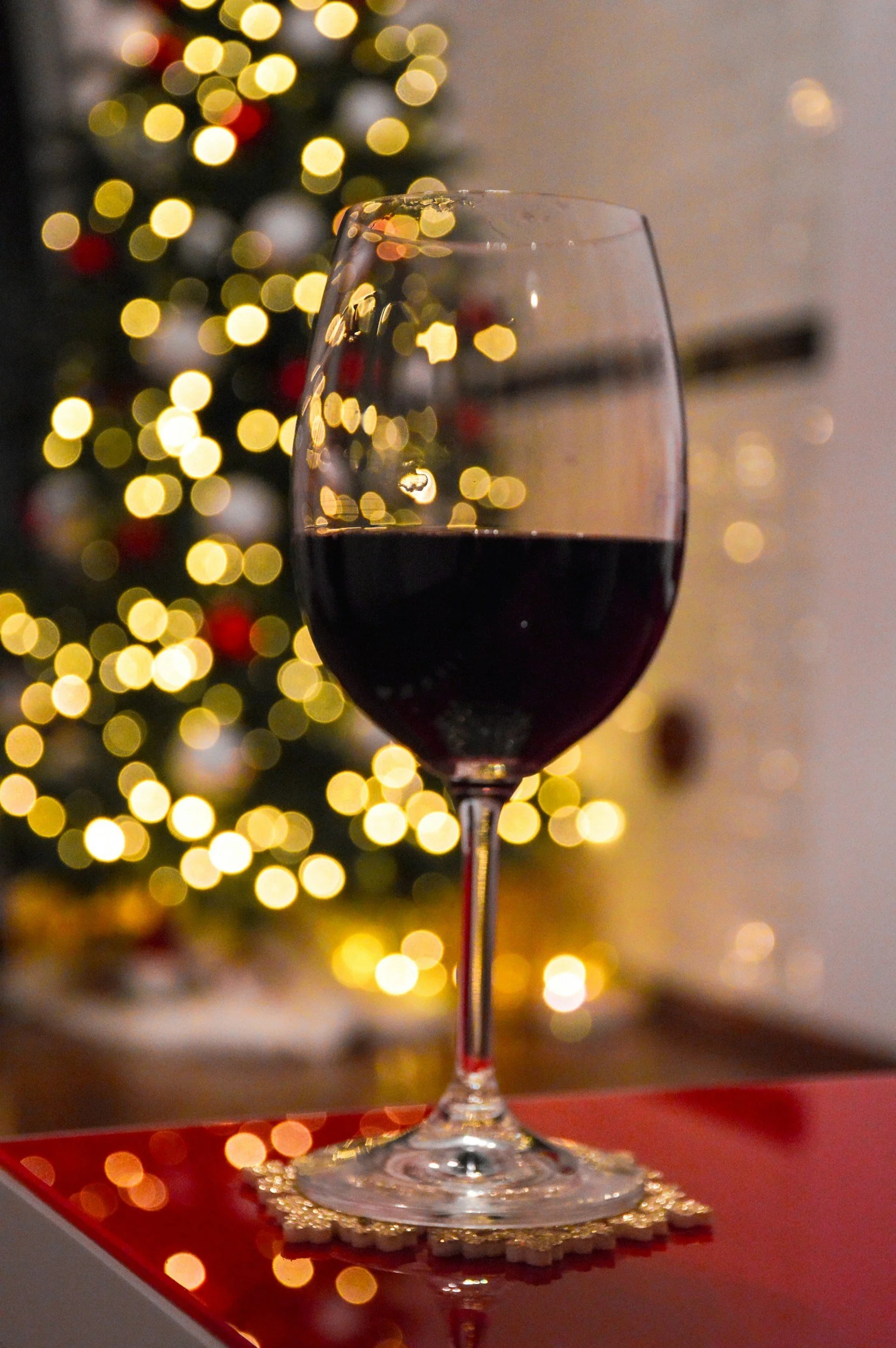 A glass of red wine on a table with a Christmas tree in the background decorated with lights and ornaments.