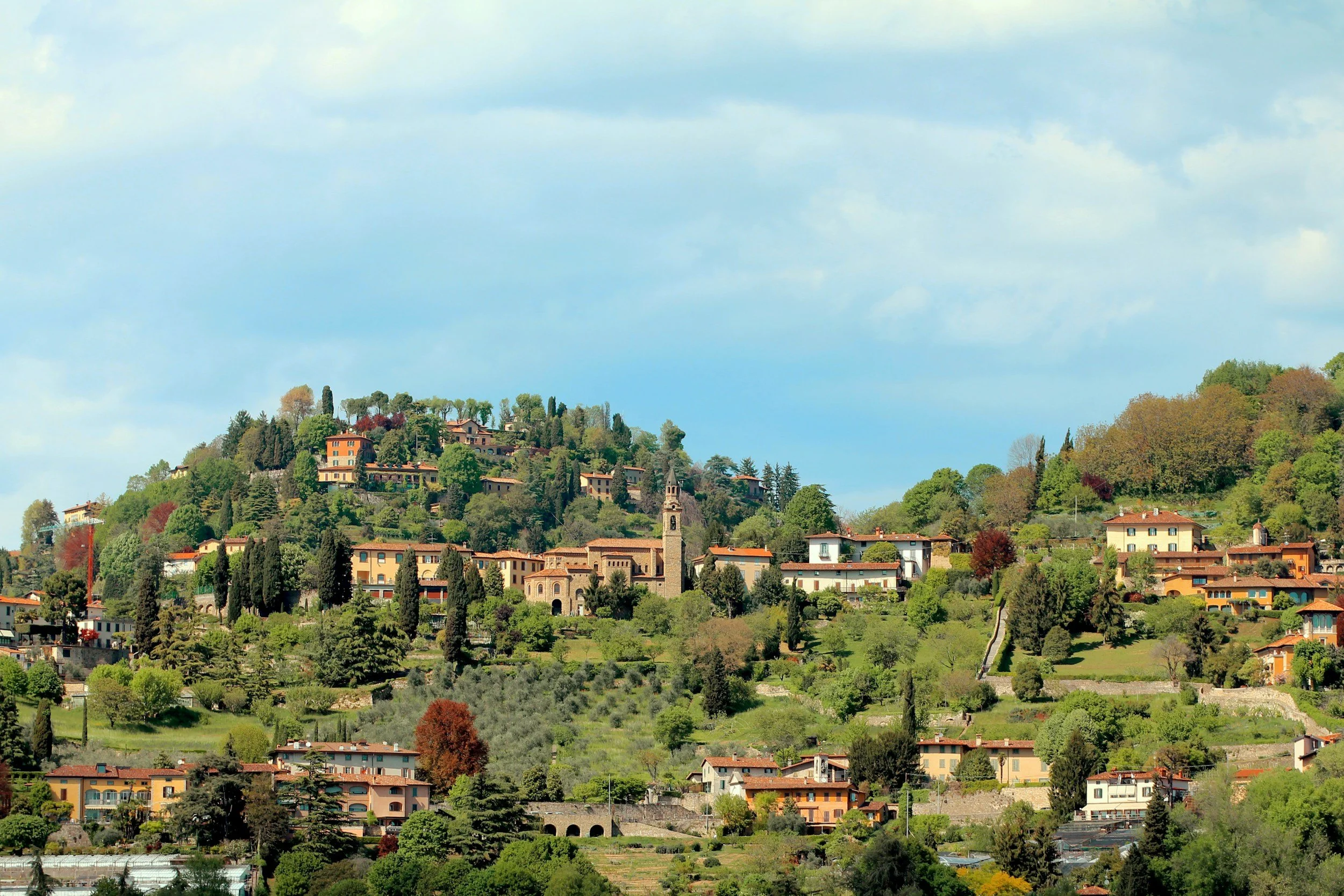 Hilly landscape with numerous colorful houses and lush green trees, topped by a partly cloudy sky.