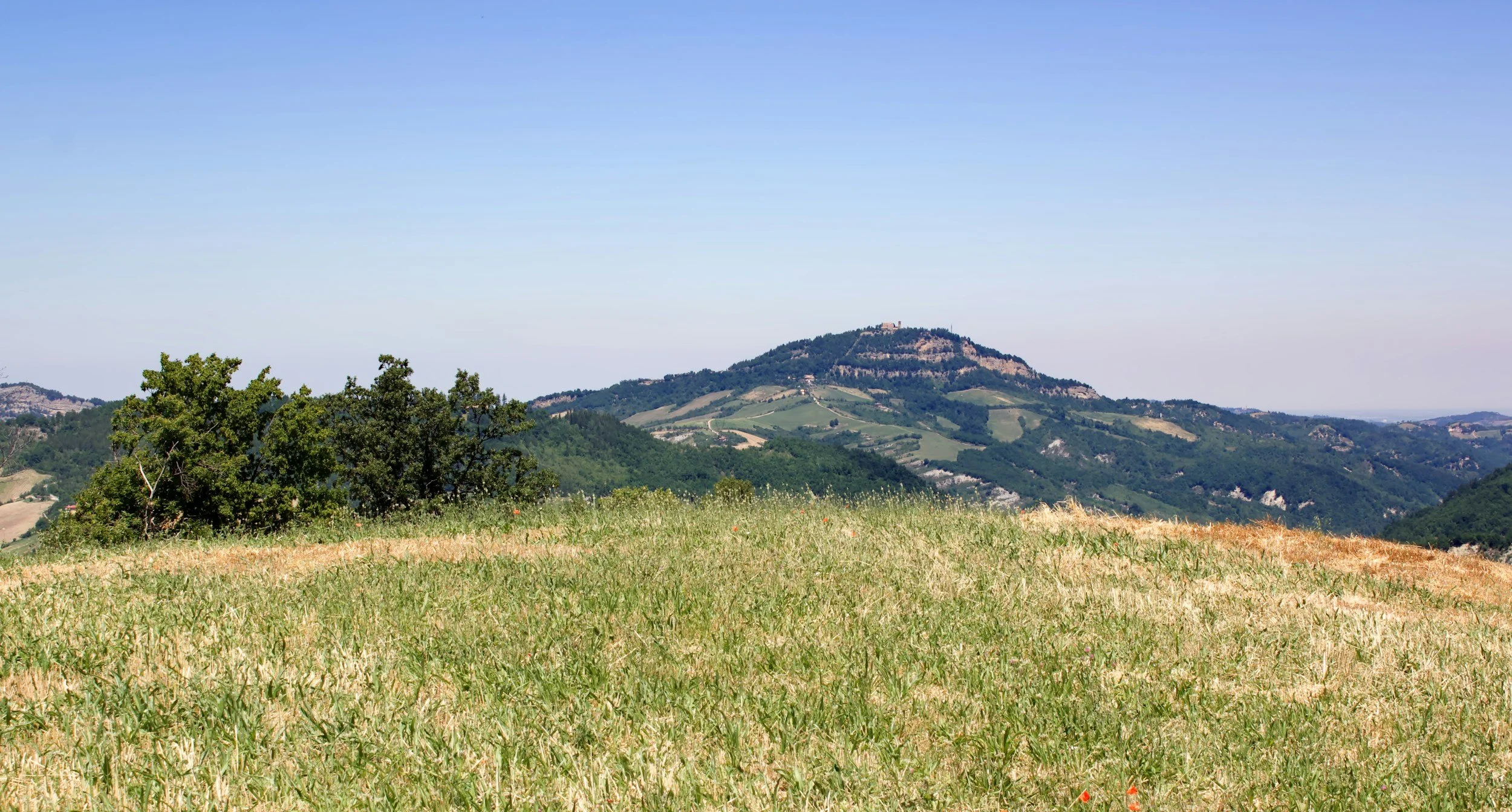 Scenic view of rolling hills with green vegetation, a mountain with a structure at the top, and a clear blue sky.
