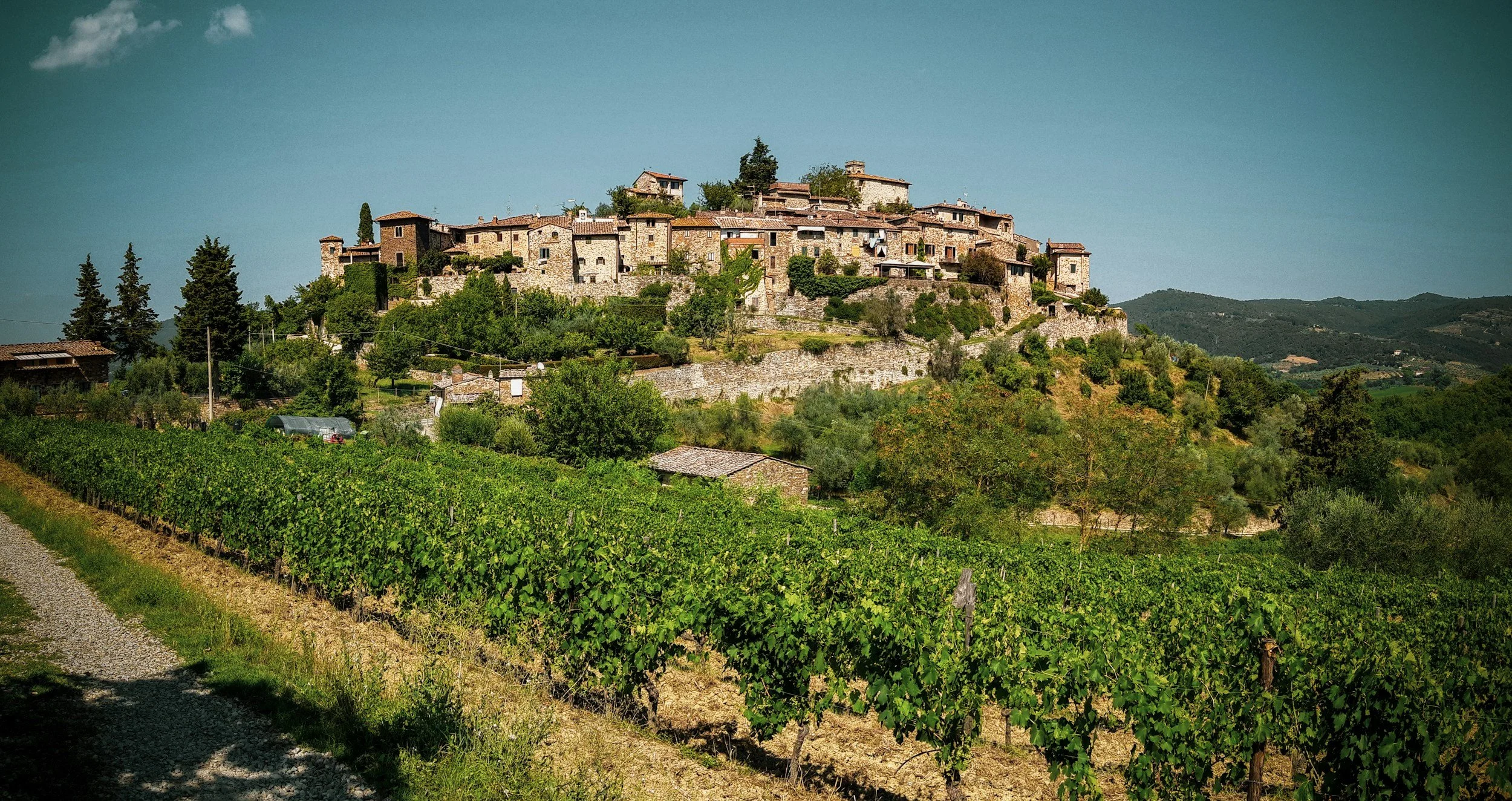 A hilltop village with stone houses surrounded by lush greenery, vineyards, and rolling hills under a clear blue sky.