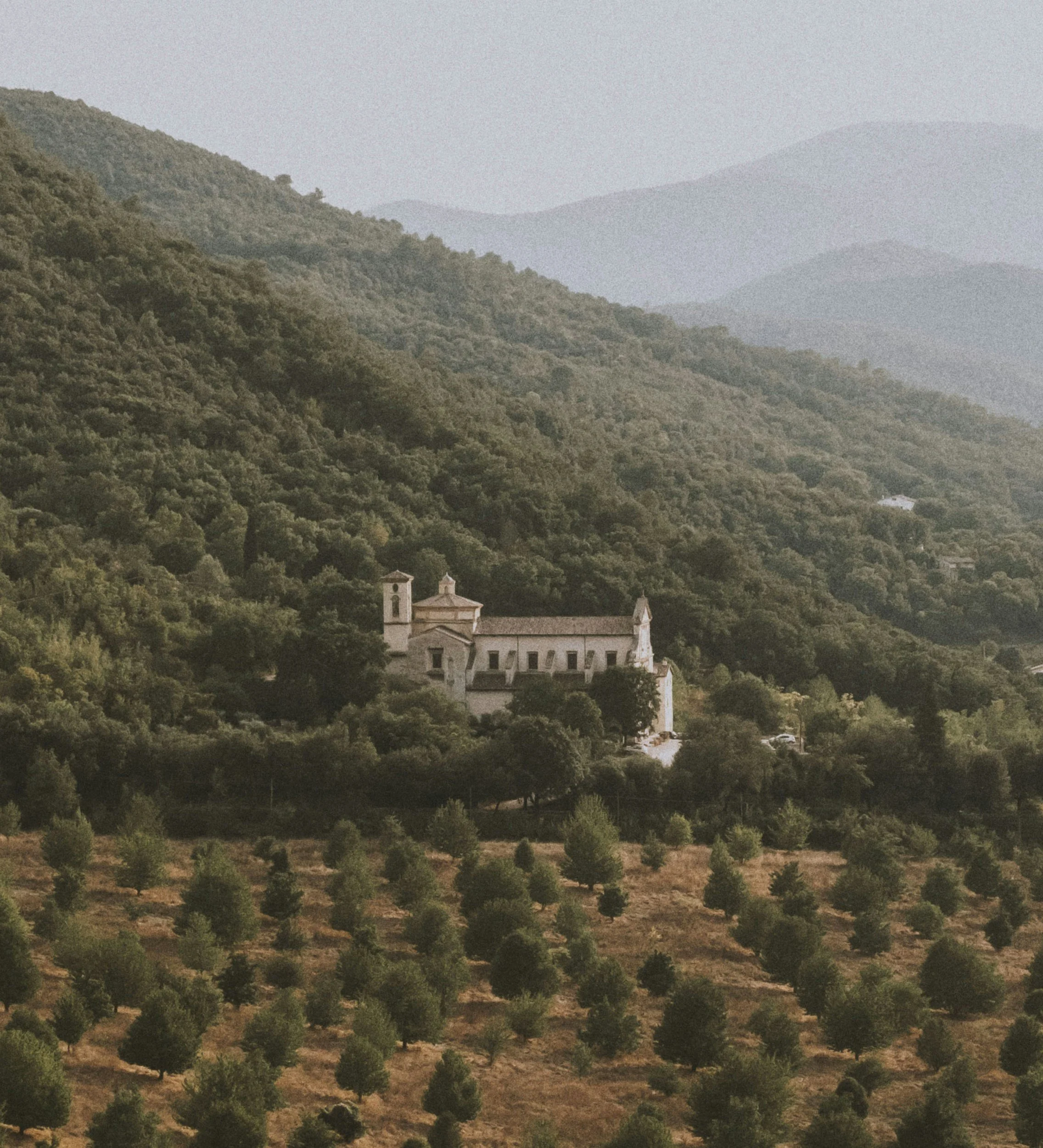 A hillside church with white walls and a bell tower, surrounded by green trees, set against a backdrop of rolling mountains.