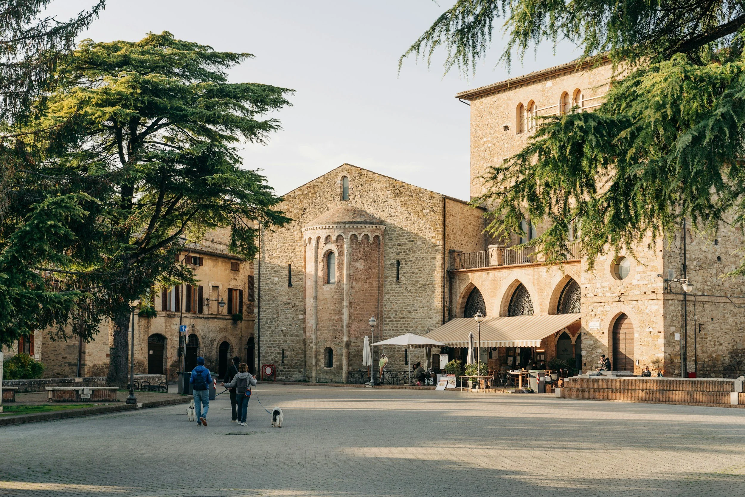 People walking with dogs in a town square surrounded by historic stone buildings and green trees, with outdoor seating and umbrellas.