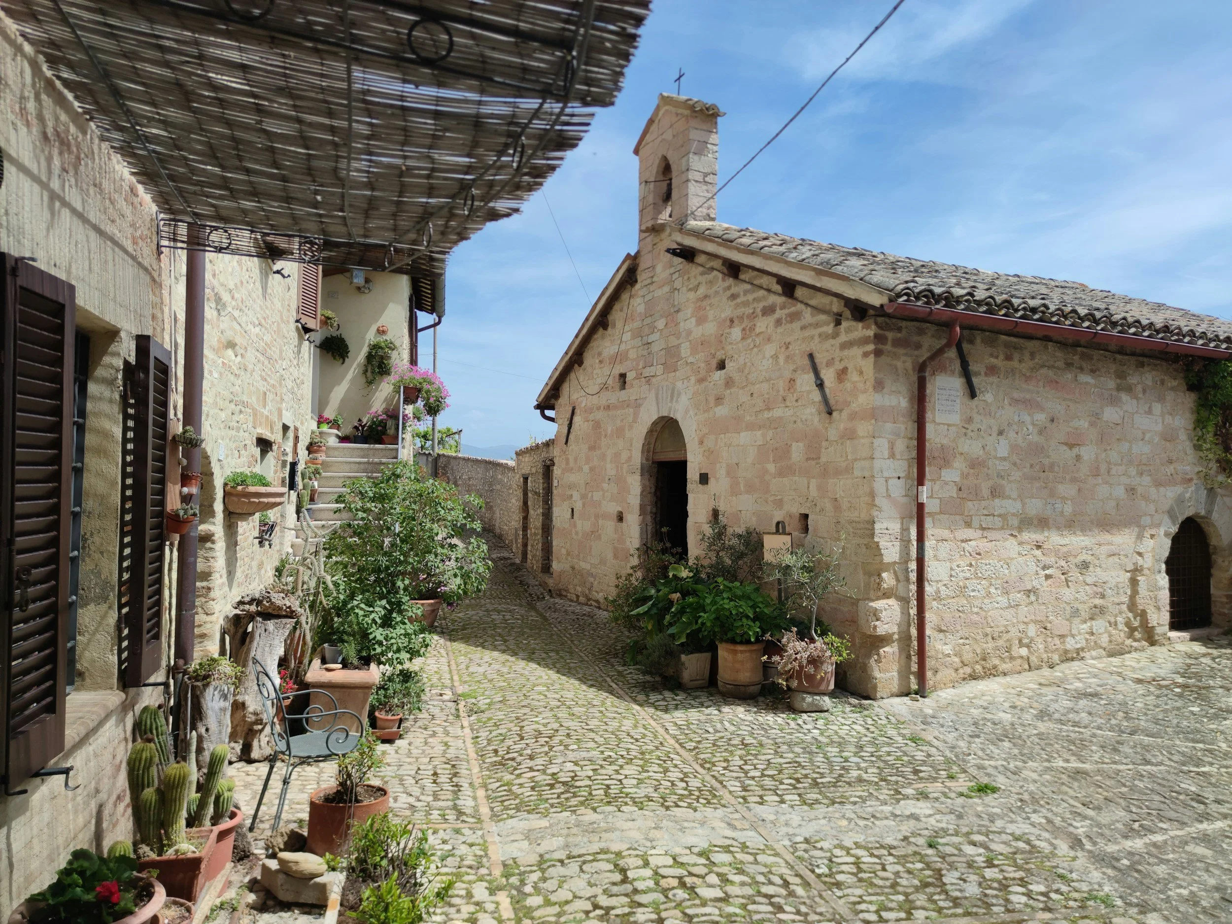 A narrow cobblestone street in a rustic village with stone buildings, potted plants, and a small church with a bell tower and cross under a blue sky.