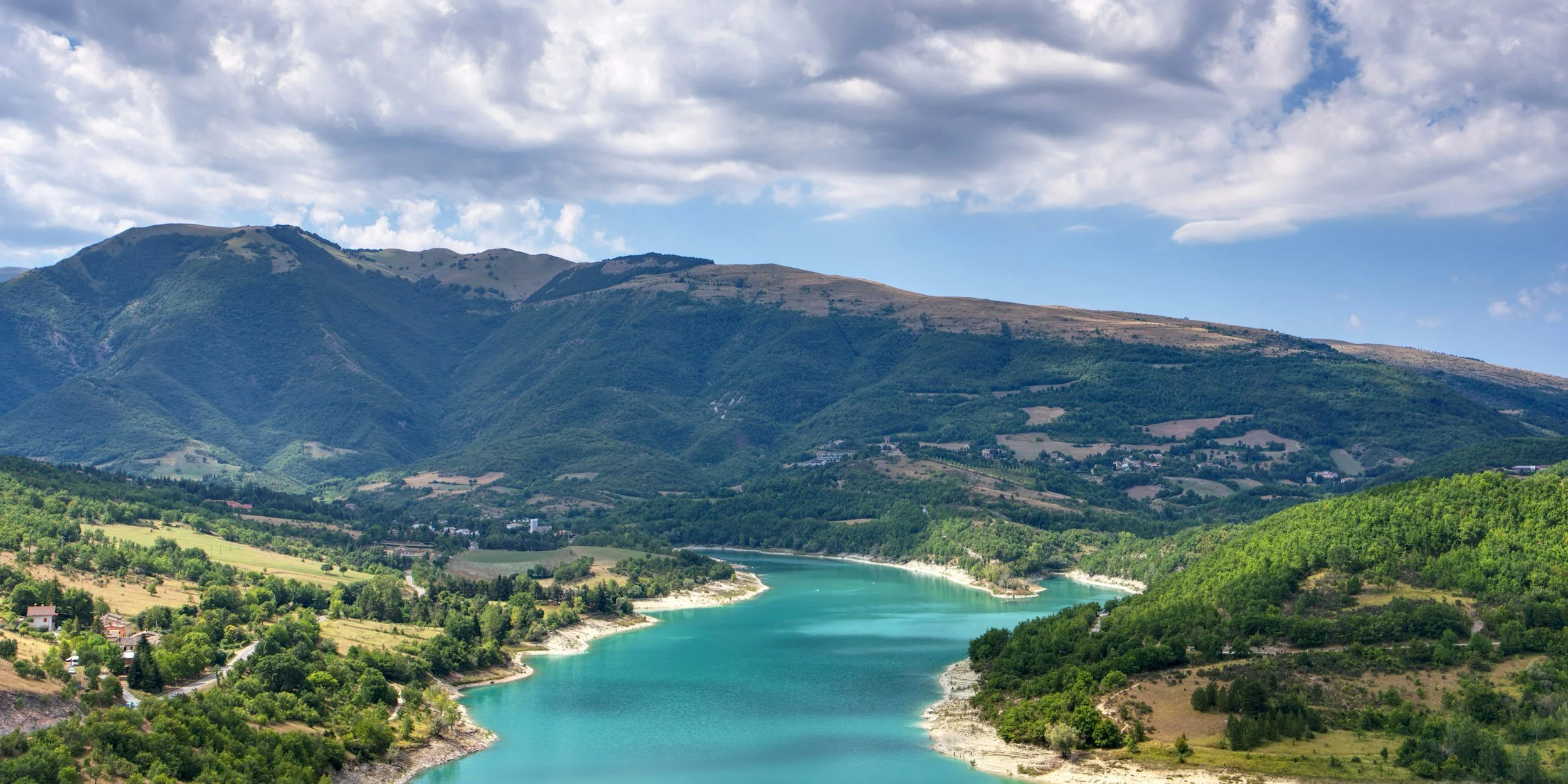 A scenic landscape with a winding river flowing through lush green forests, with mountains and hills in the background under a partly cloudy sky.