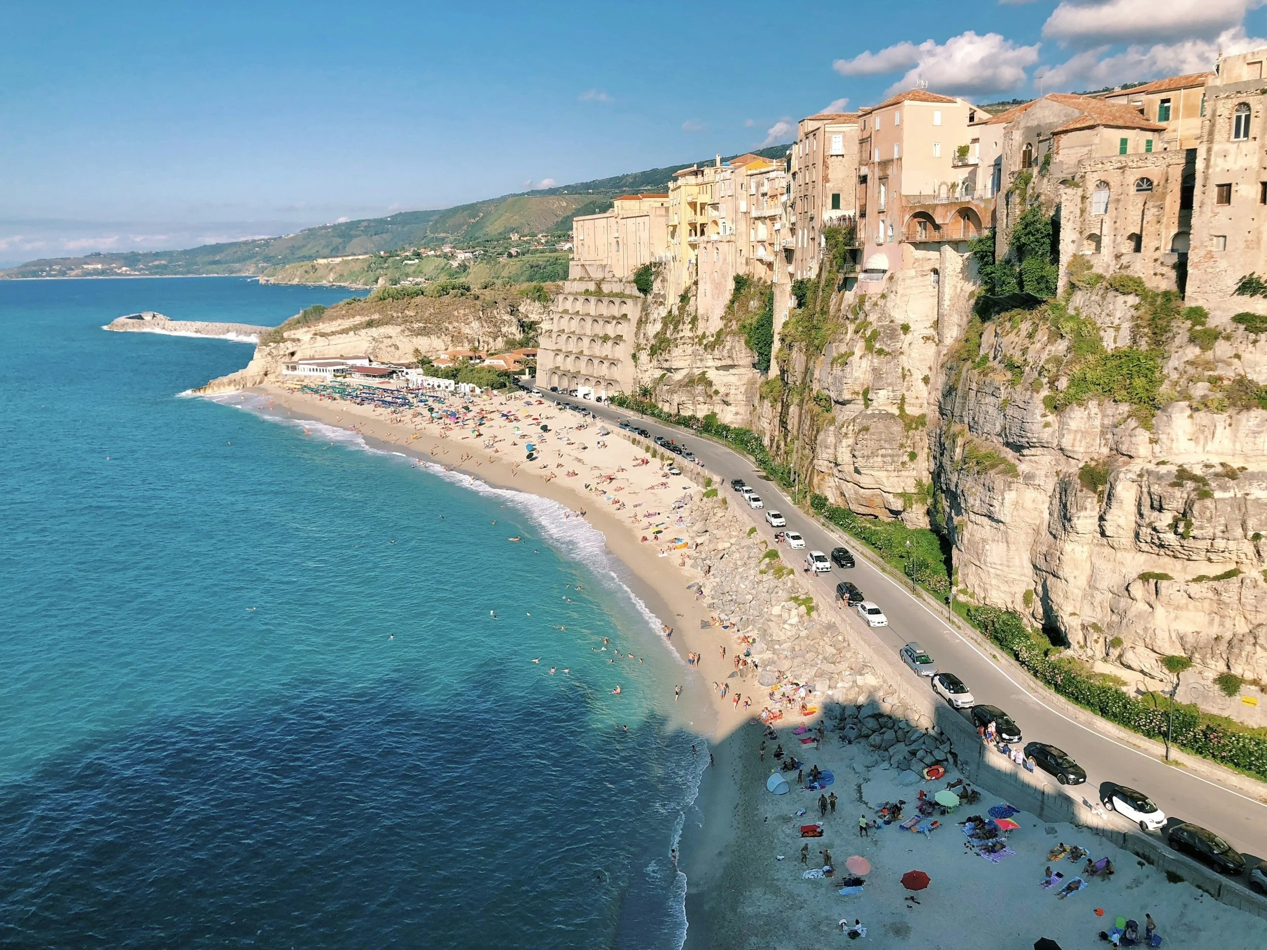 A coastal scene with a sandy beach filled with colorful umbrellas and people enjoying the shore. The beach is adjacent to a road lined with parked cars, with a steep cliff covered in green vegetation and pastel-colored buildings overlooking the sea. The water is clear and blue, with gentle waves lapping the shore. In the background, there are rolling hills under a partly cloudy sky.