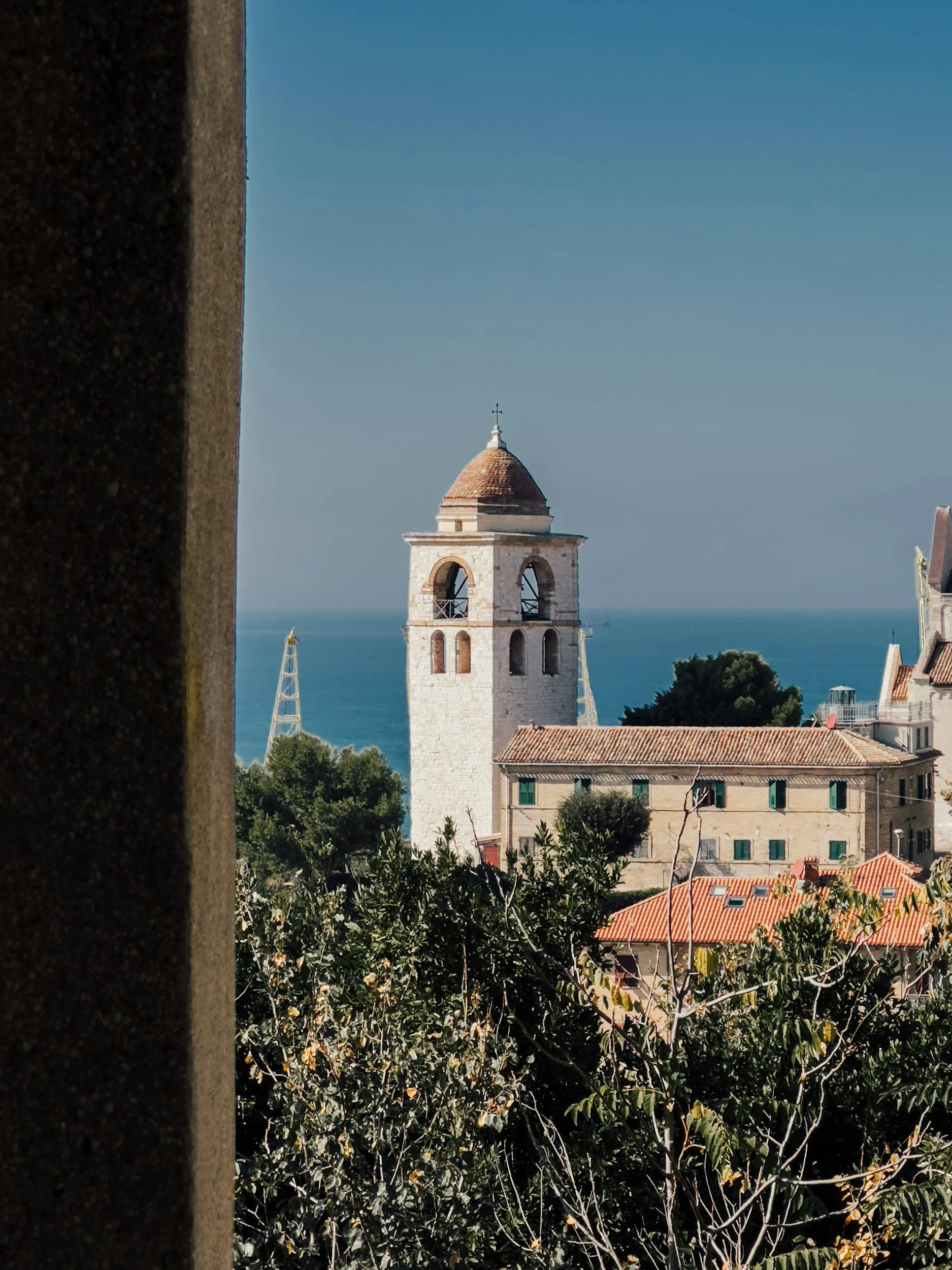 View of a historic church tower with a cross on top, surrounded by trees and red-tiled roofs, with the sea in the background, partly obscured by a stone wall and foliage.