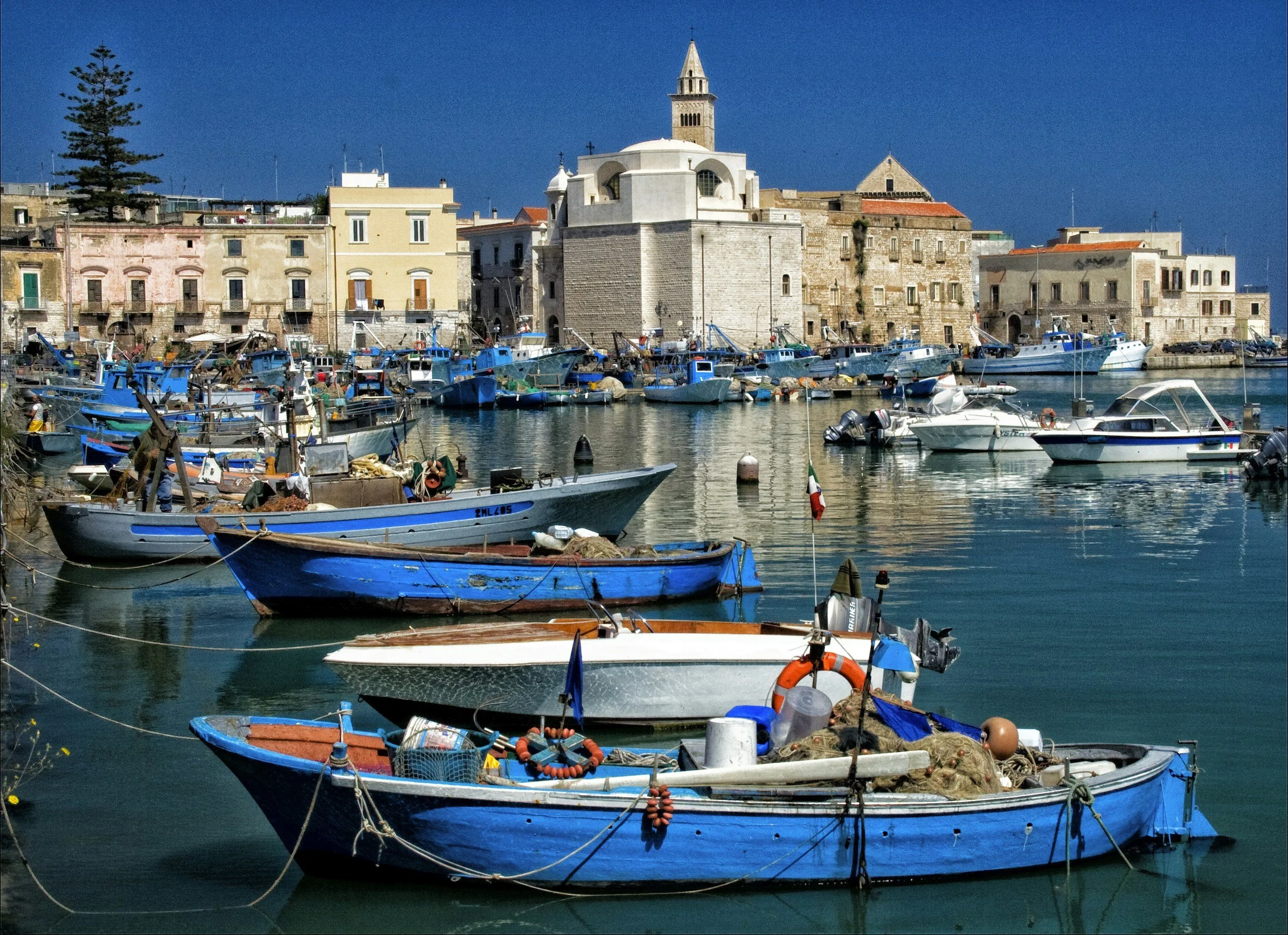 A harbor with several small boats docked, with a backdrop of historic buildings and a church steeple, under a clear blue sky.