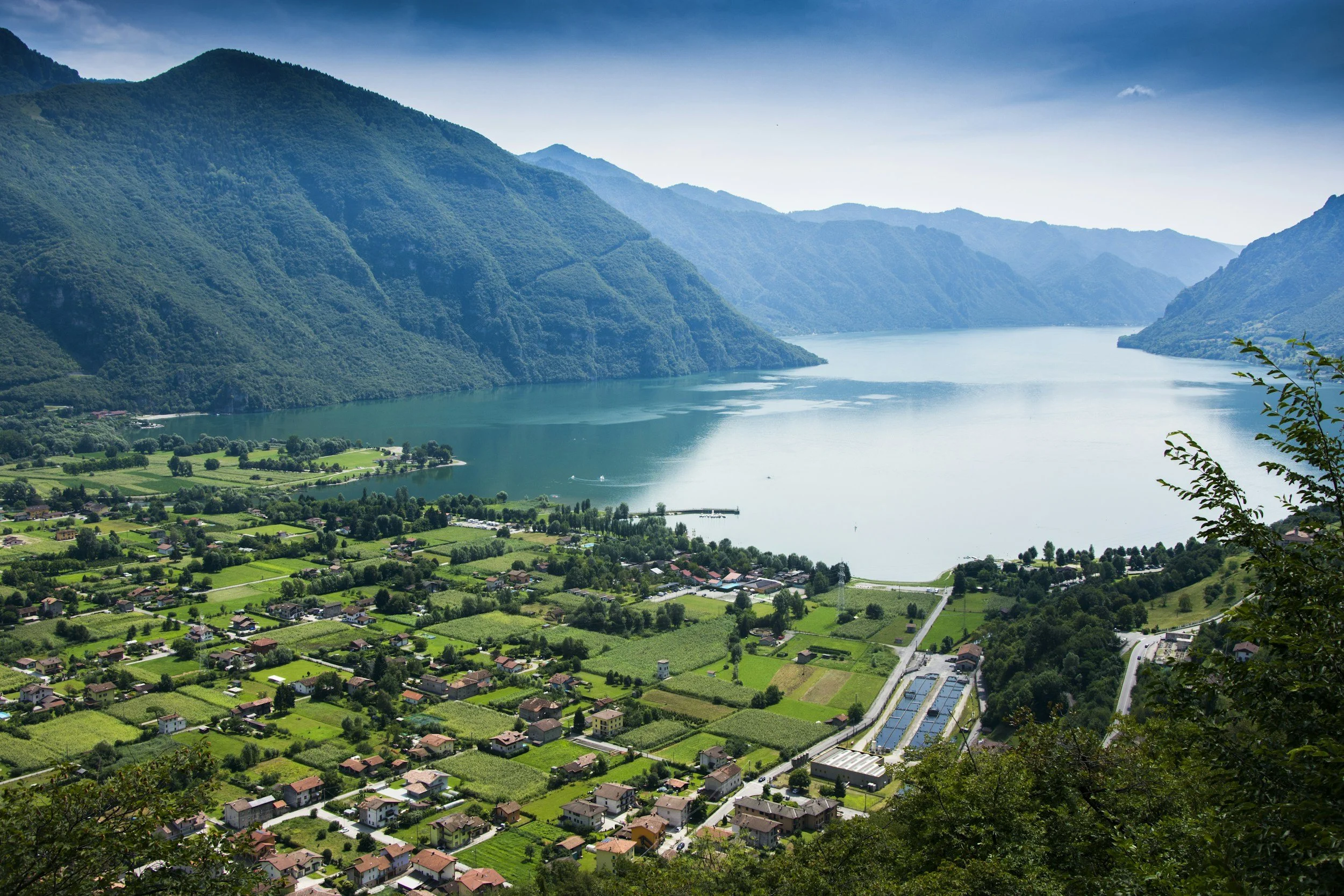 A scenic view of a river winding through lush green fields and small houses, surrounded by tall, forested mountains under a blue sky.