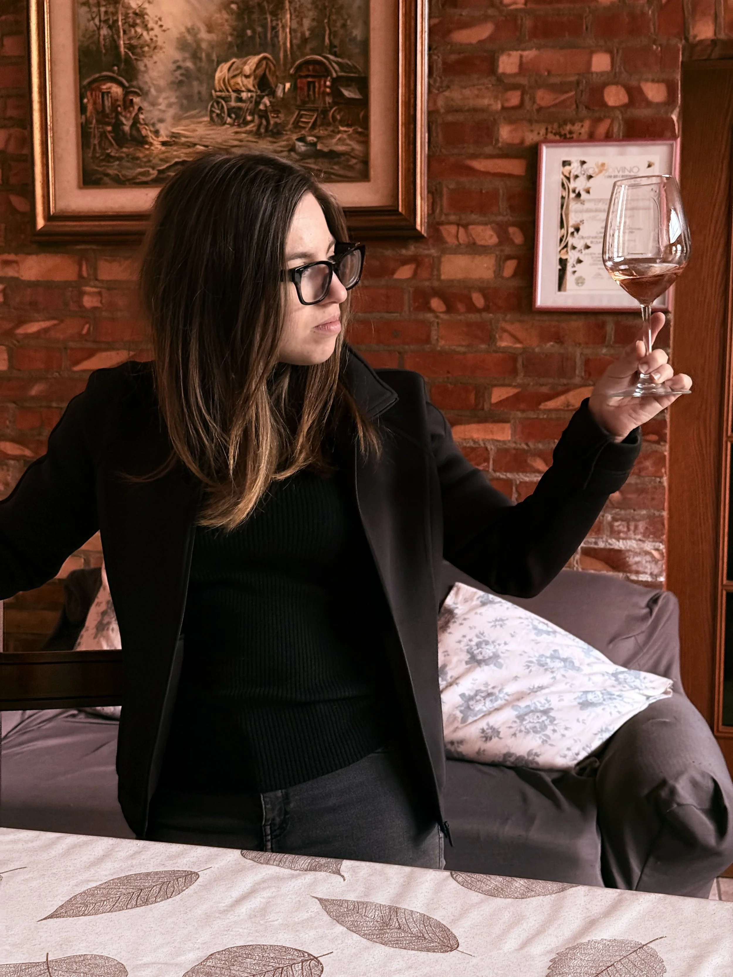 A woman with glasses and long brown hair, wearing a black jacket and black shirt, holding a glass of wine, sitting in a room with brick walls, framed pictures, and a bed with floral bedding.