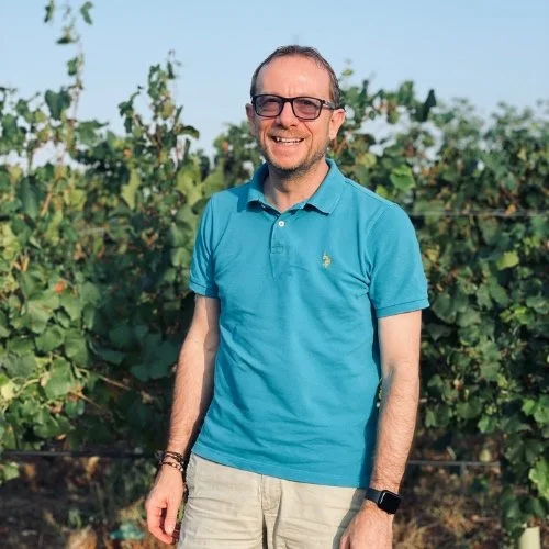 Man wearing glasses and a blue polo shirt smiling outdoors in a vineyard.