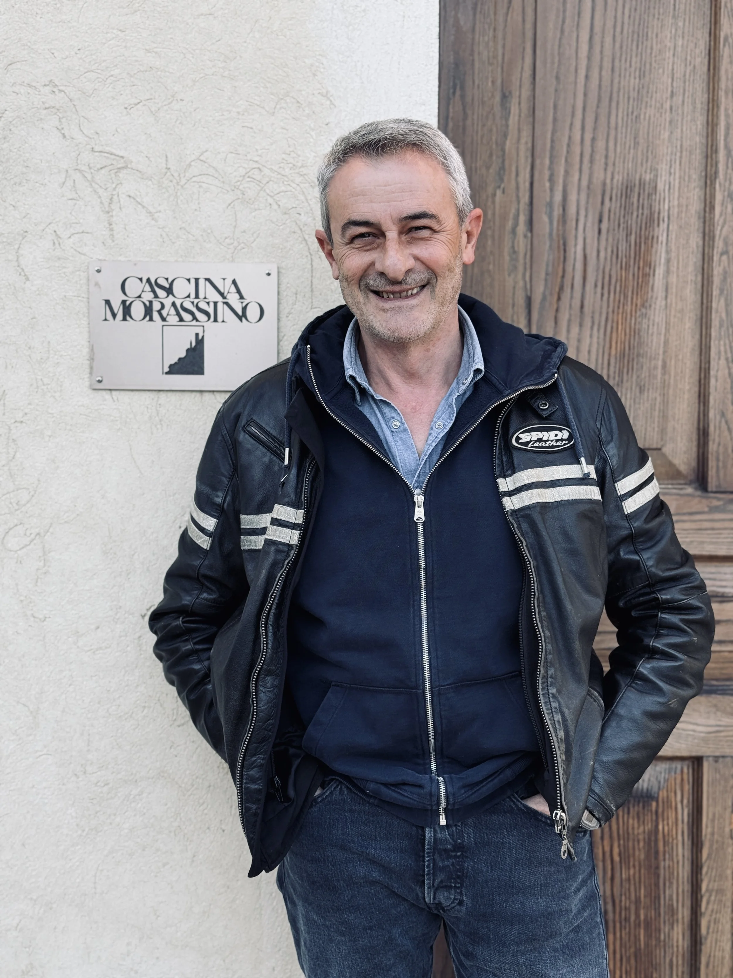A smiling middle-aged man with short gray hair and facial stubble, wearing a leather jacket over a light blue zip-up hoodie, standing in front of a textured wall with a sign that reads "Cascina Morassino" and a wooden structure.