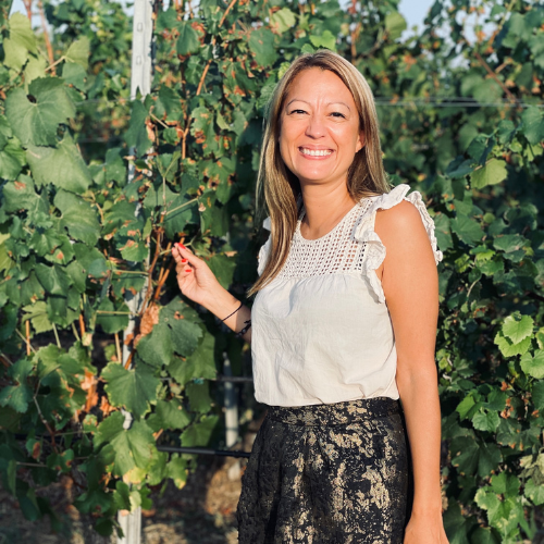 Woman smiling in a vineyard, holding a vine with green leaves.