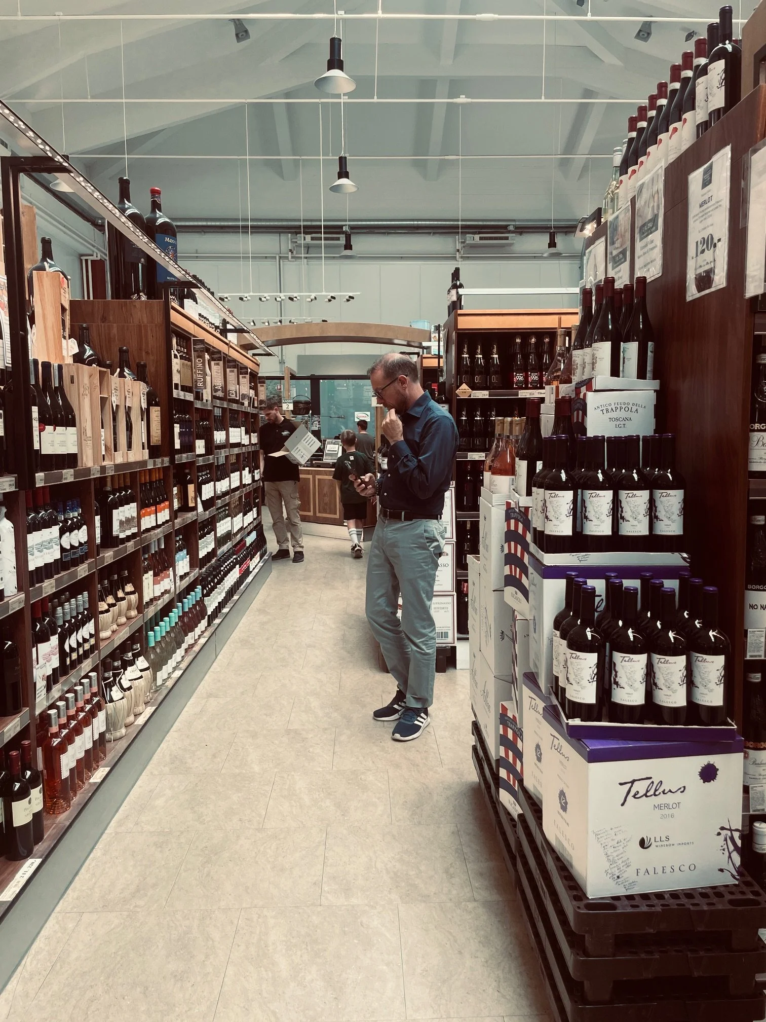 Inside a wine store with a man in a blue shirt and gray pants looking at his phone, surrounded by shelves stocked with wine bottles.
