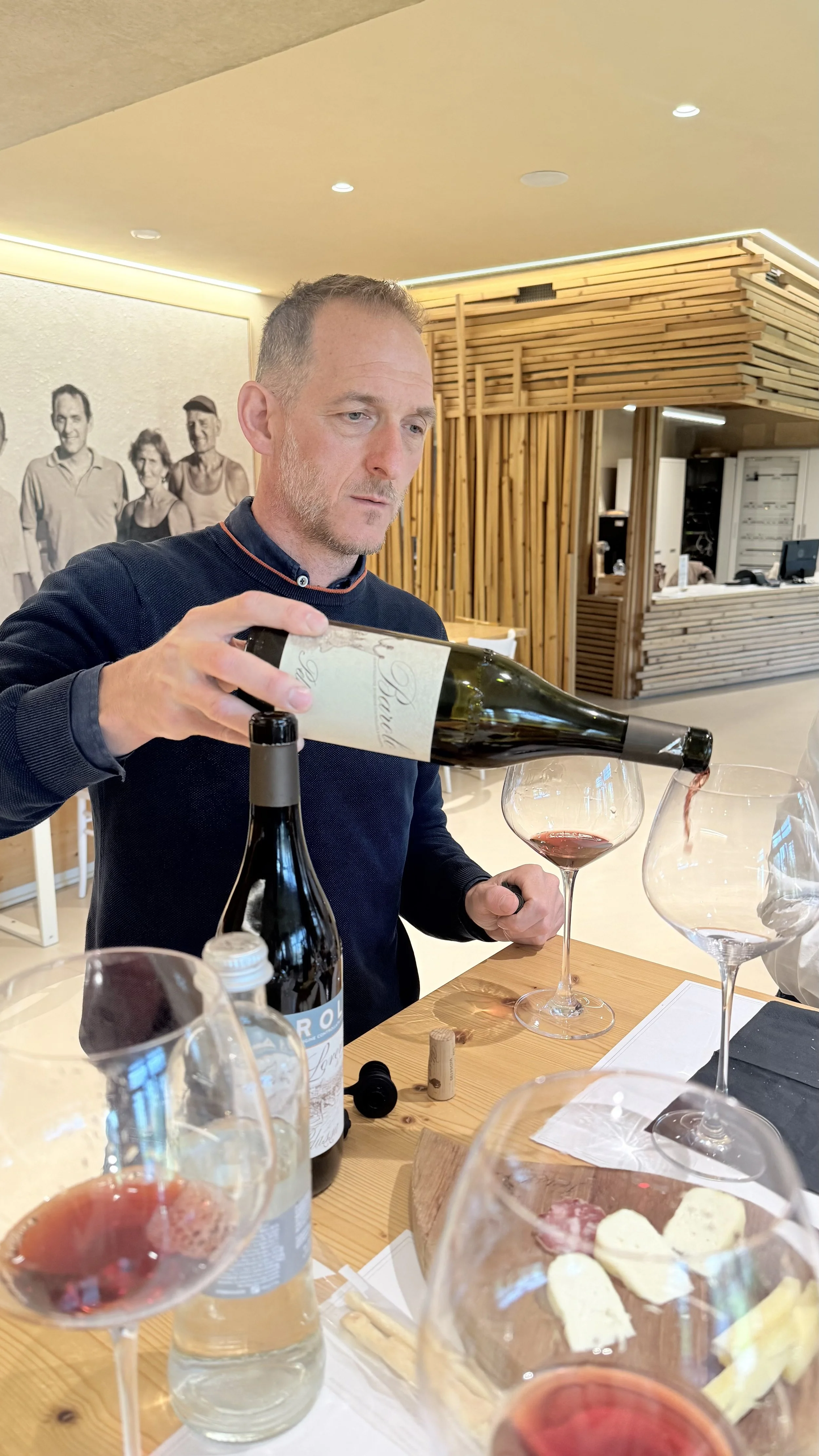 A man pouring red wine into a glass at a table with bottles, glasses, and cheese, in a modern restaurant with wooden decor.
