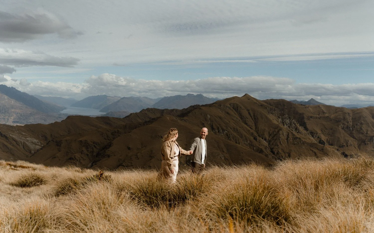 Bride and groom walking in the tussock at Vanguard Peak. Lake Wakatipu and the Remarkables in the background.
