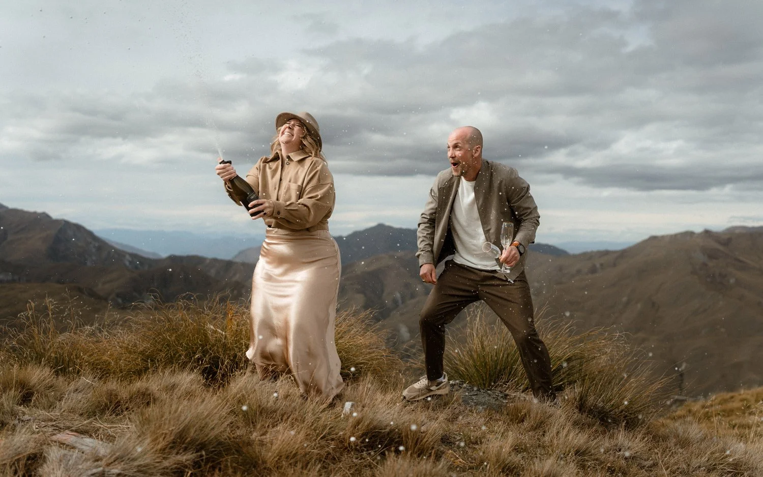 Queenstown wedding couple popping Central Otago champagne during their Vanguard Peak heli elopement. New Zealand mountains in the background. 
