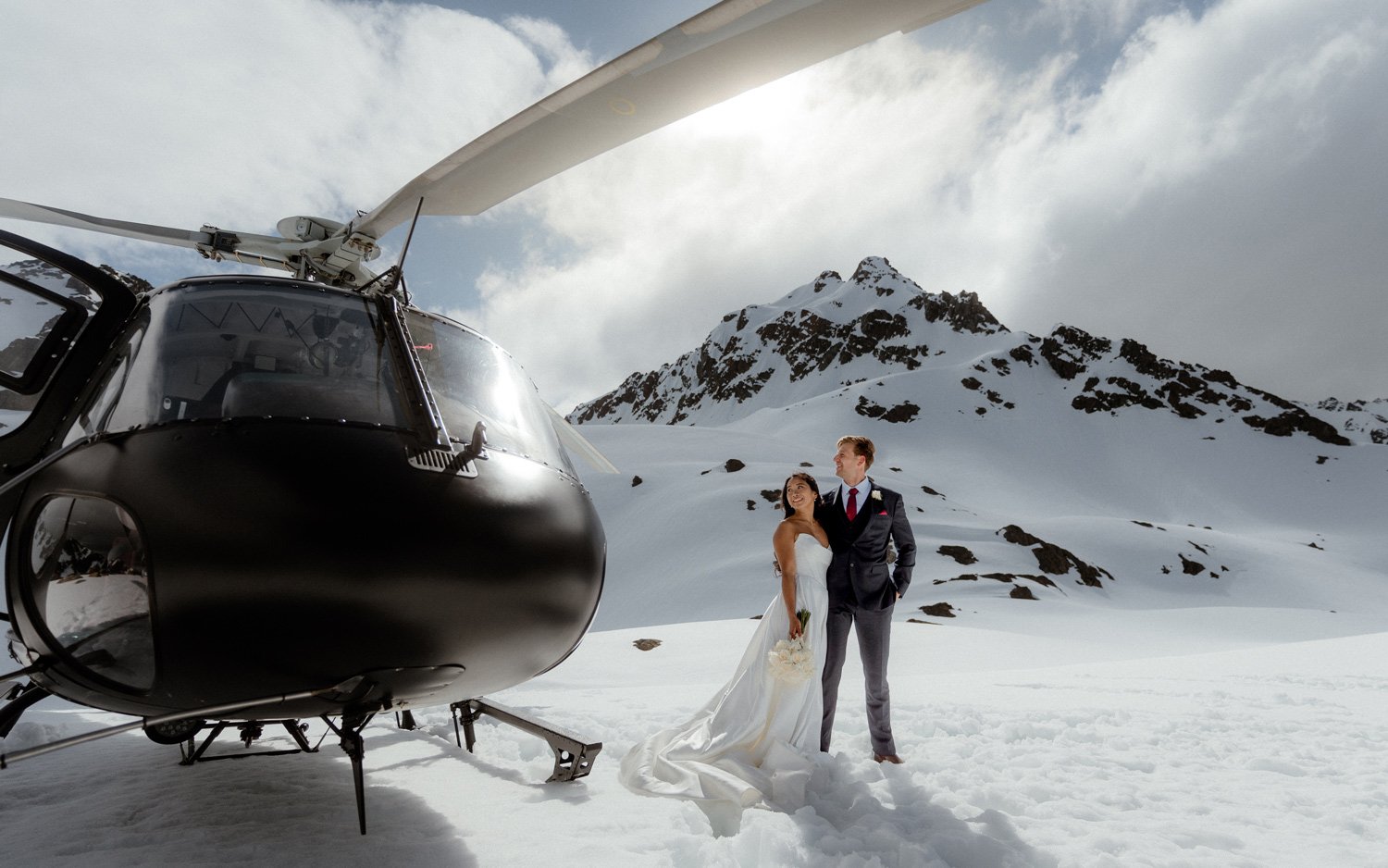 heli wedding at The Humboldt's mountains in Queenstown. Bride & groom standing in the snow by Heliworks helicopter at their mountain wedding in Queenstown. 