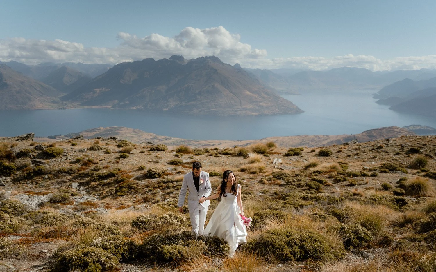 The Remarkables heli elopement location. Couple walking with The Remarkables, Lake Wakatipu and Queenstown in the background. Elopement photographer Ricky B Good Photography