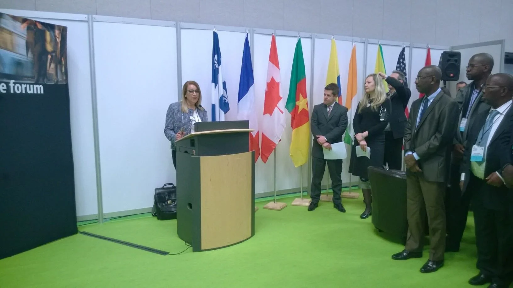 A woman is giving a speech at a podium during a conference, with multiple international flags displayed behind her and an audience of diverse individuals listening attentively.