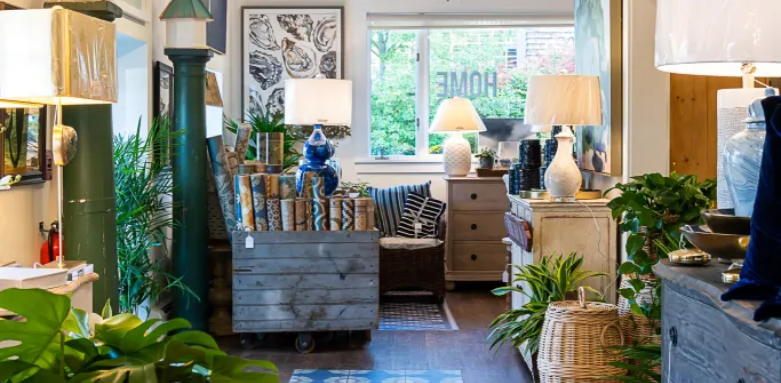 Cozy living room with assorted furniture, green plants, table lamps, and decorative items, overlooking a window with greenery outside.