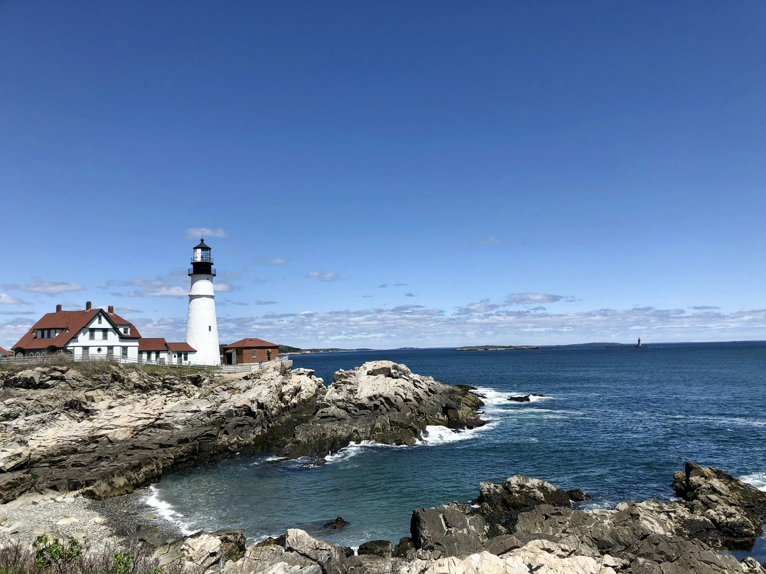 A lighthouse on rocky coastline next to a house with a red roof under a blue sky with scattered clouds, overlooking the ocean.