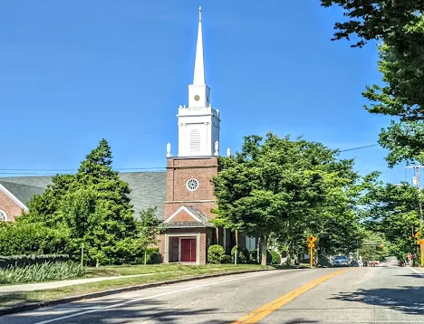 A church with a white steeple and brick facade, surrounded by green trees, on a sunny day with a clear blue sky.