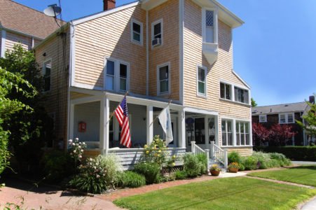A multi-story house with a front porch, American flag, and a garden with bushes and flowers, under a clear blue sky.
