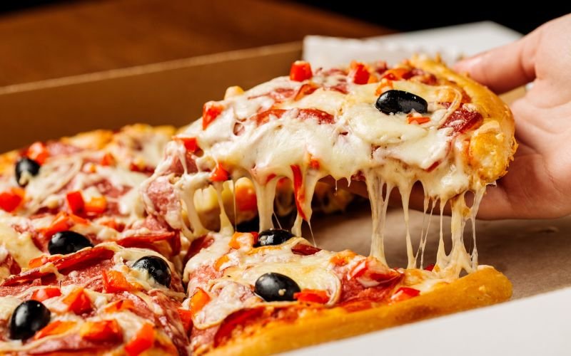 Slice of pizza with cheese, black olives, red peppers, and ham being served from a pizza box.